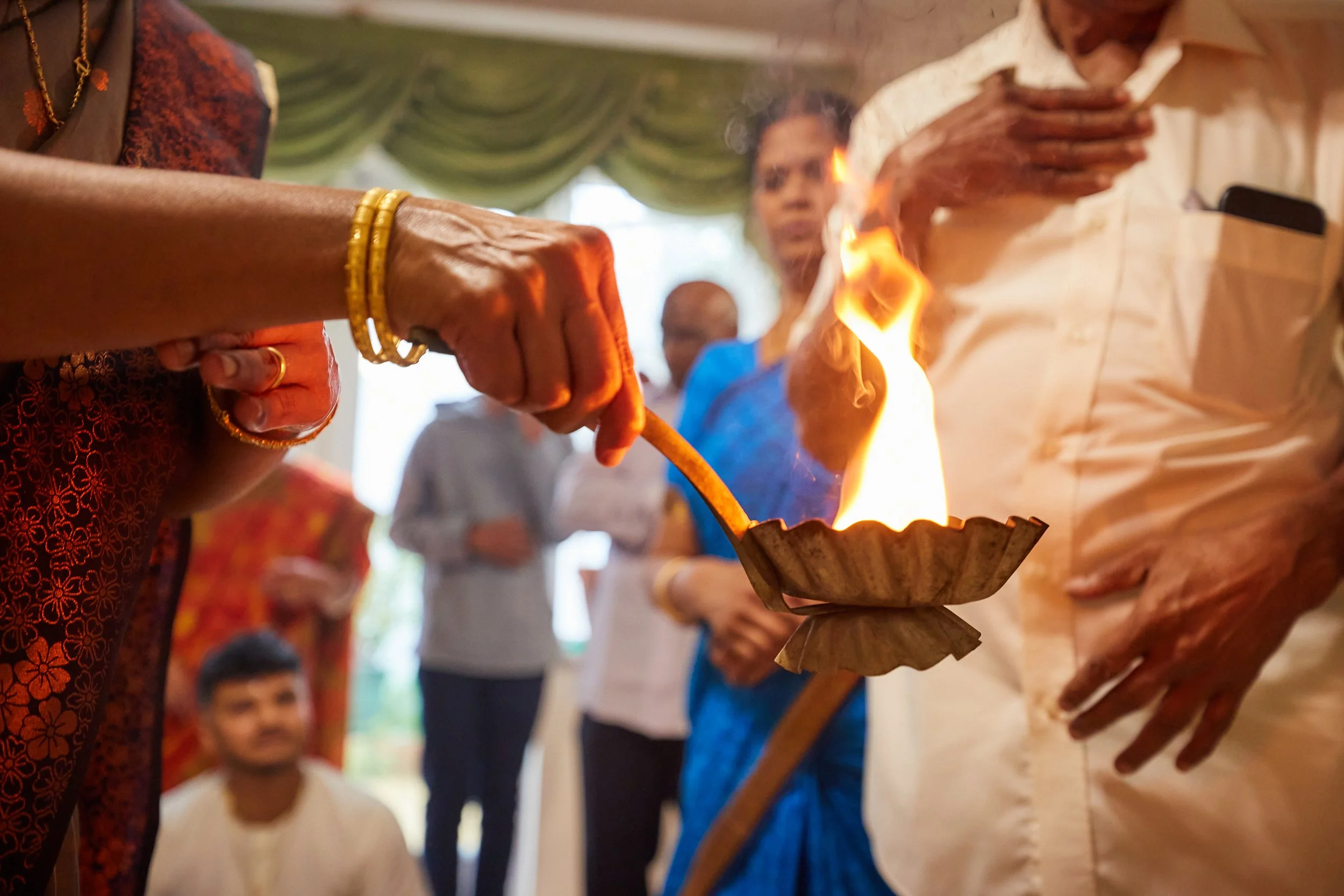An Indian woman in traditional attire holds a lit oil lamp during a ceremony, with other people observing in the background.