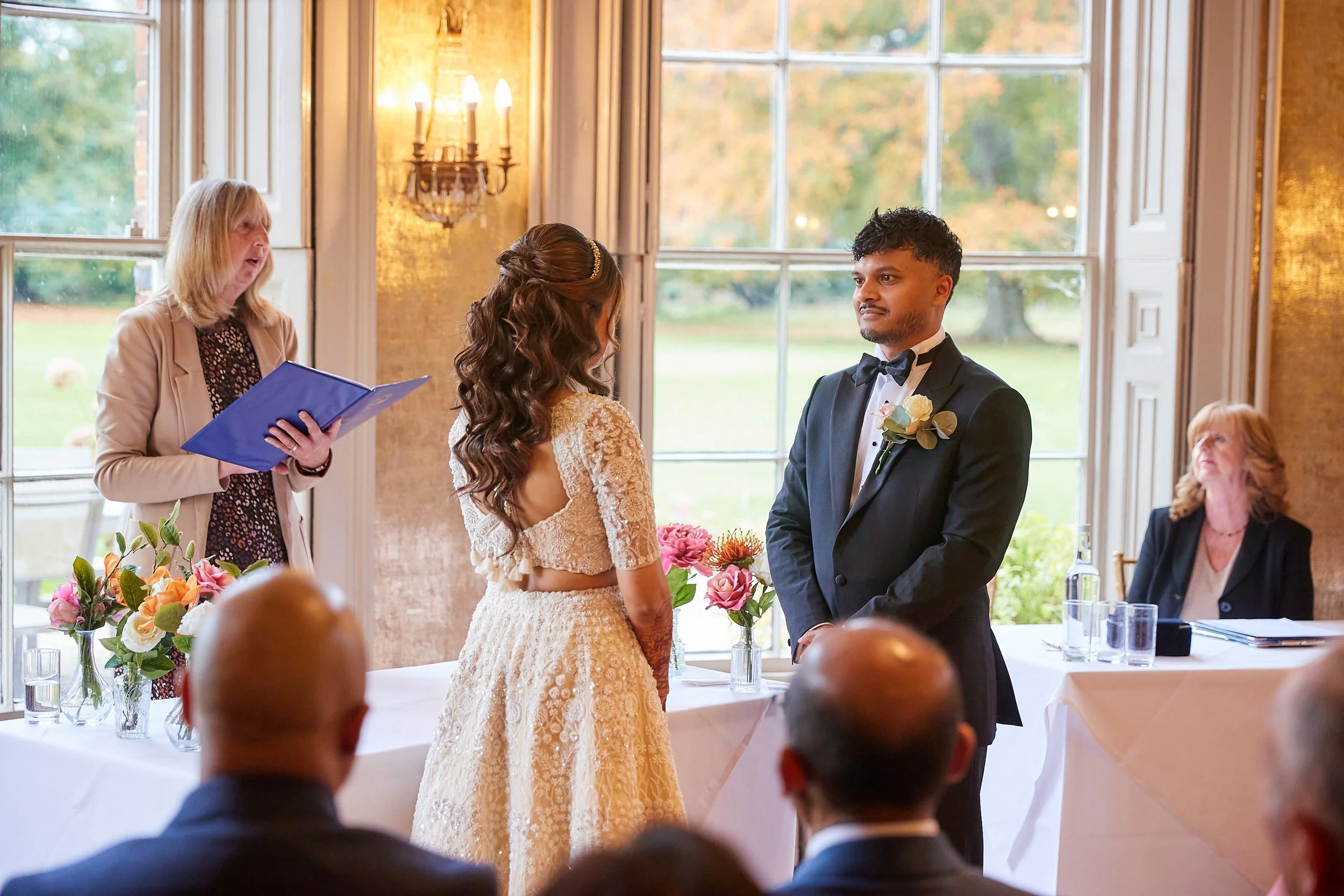 A bride and groom standing face to face during their wedding ceremony in a room with large windows, with an officiant woman holding a blue folder in the background and seated guests in the foreground.