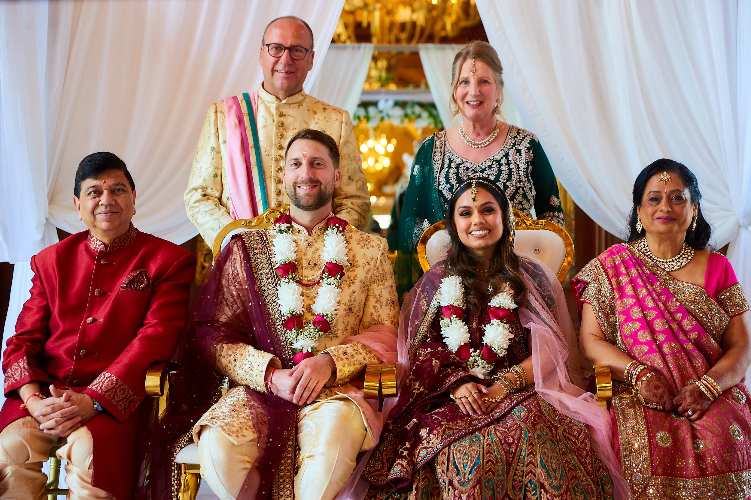 Group of seven people celebrating a wedding, with a bride and groom in the center, all dressed in traditional Indian attire, sitting and standing in front of a white cloth backdrop with drapes.