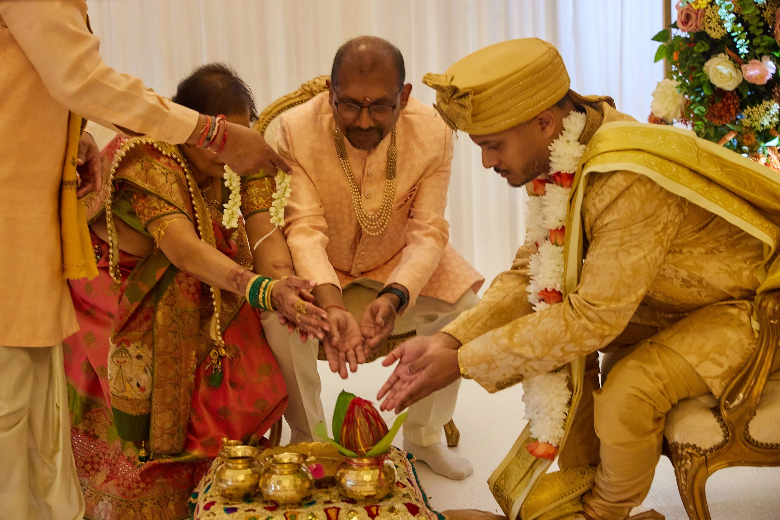 Indian couple and family participating in a traditional wedding ritual, with woman in colorful saree and gold jewelry, man in gold patterned traditional attire, family members assisting, and decorative elements in the background.