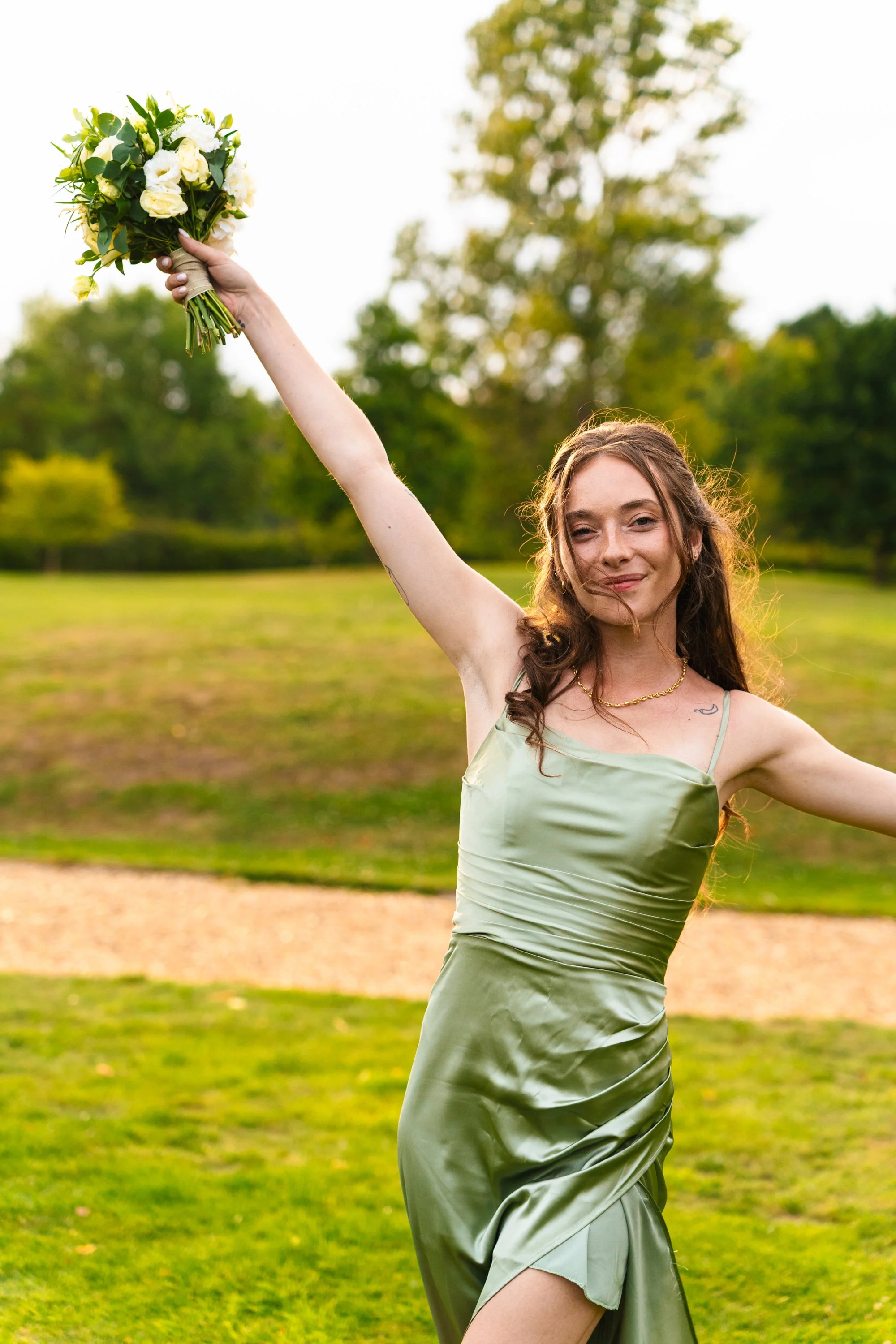 A woman in a green satin dress holding a bouquet of white and yellow flowers, smiling outdoors in a park during daytime.