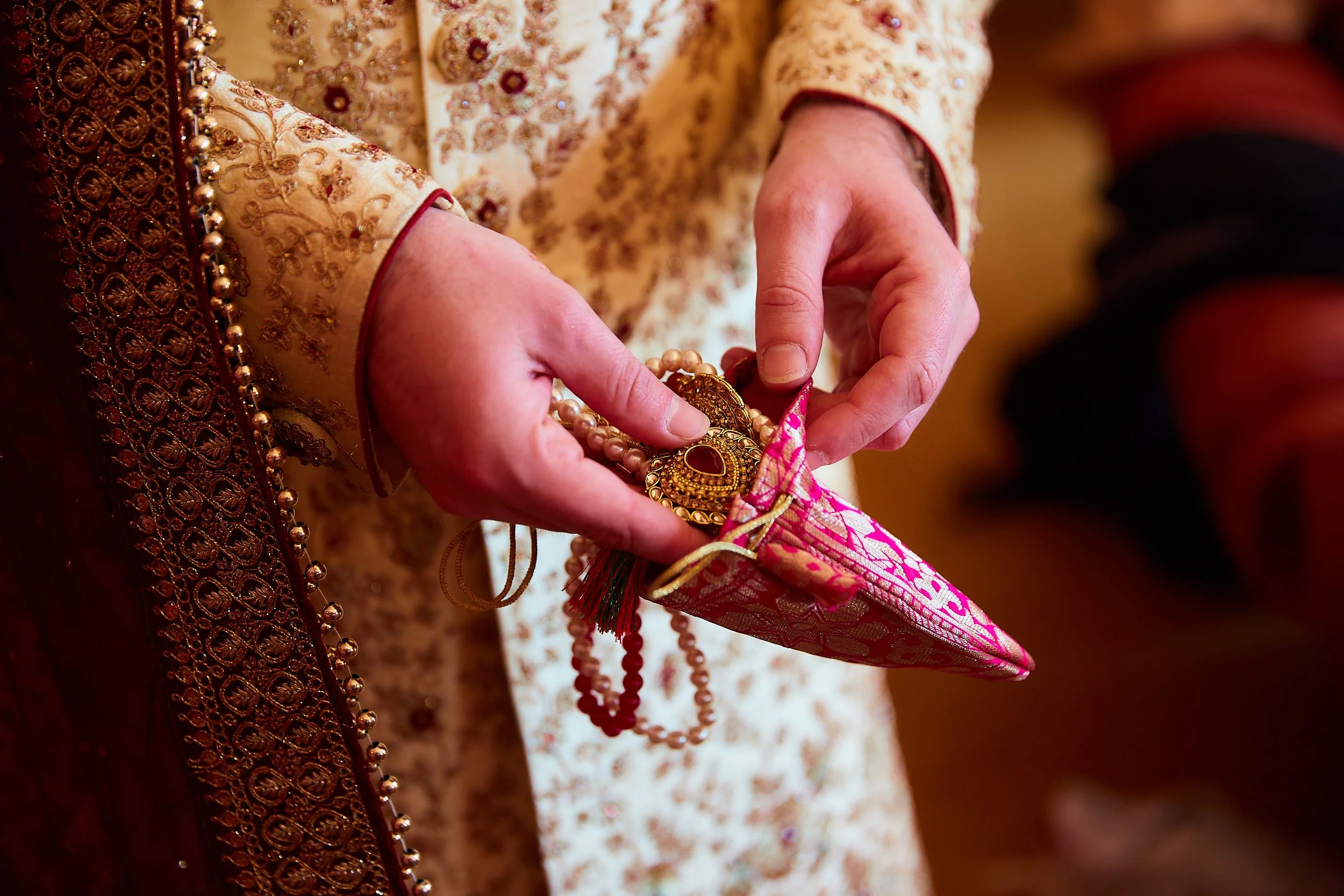 Person wearing traditional embroidered clothing holding a pink and gold embroidered pouch containing jewelry, including a gold necklace and earrings.