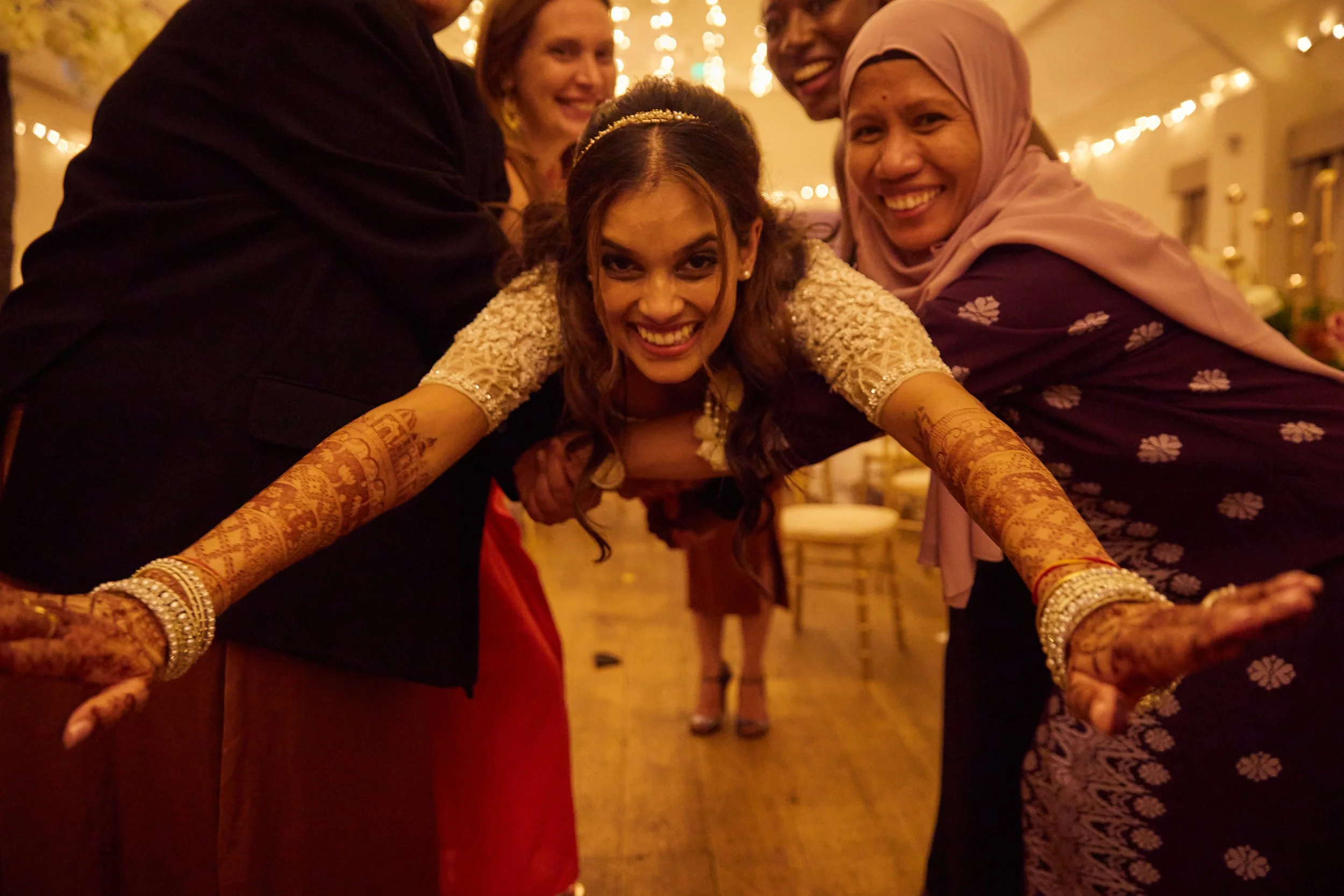 Group of women celebrating, with the central woman extending her arms forward, smiling, and wearing traditional South Asian attire and henna designs on her arms, in a festively decorated indoor setting.