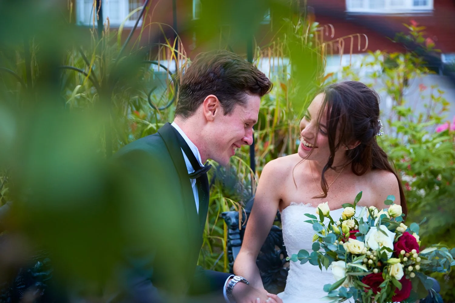 A newlywed couple smiling joyfully at each other, surrounded by greenery and flowers, during their wedding celebration.