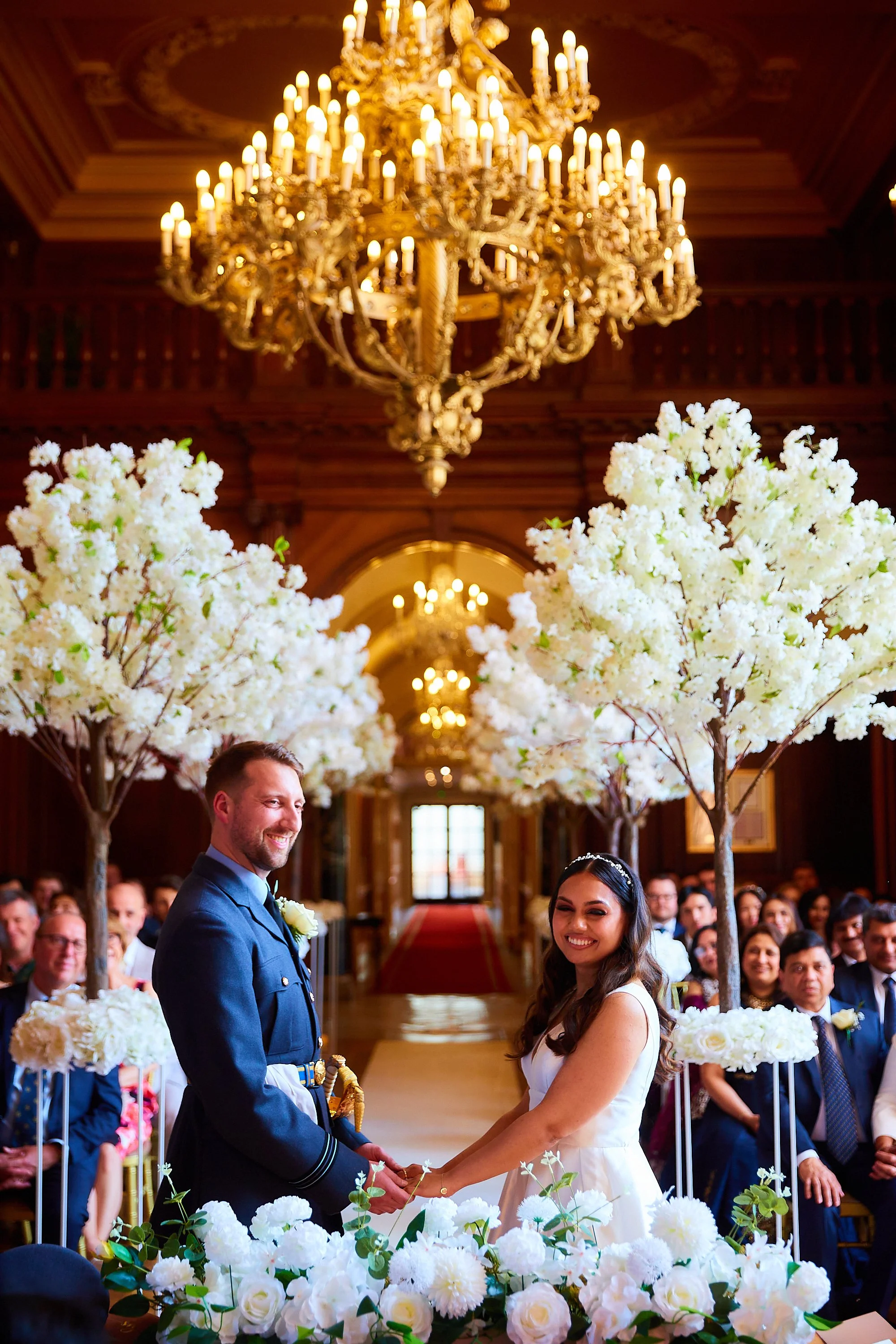 A wedding ceremony taking place in a grand hall with a chandelier, white floral trees, and an audience of guests. A bride and groom are holding hands and smiling at each other.