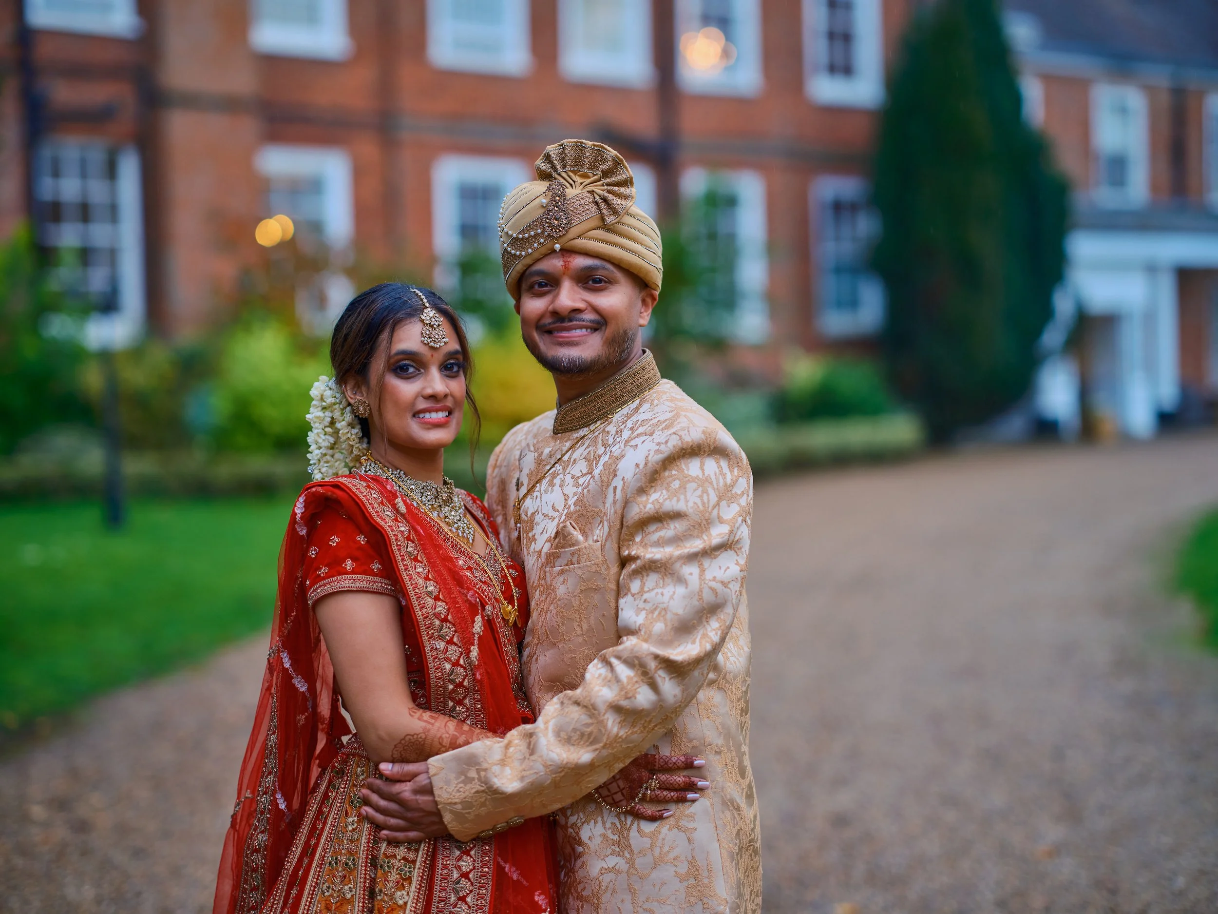 A couple dressed in traditional Indian wedding attire standing outdoors in front of a brick building and greenery, smiling at the camera.