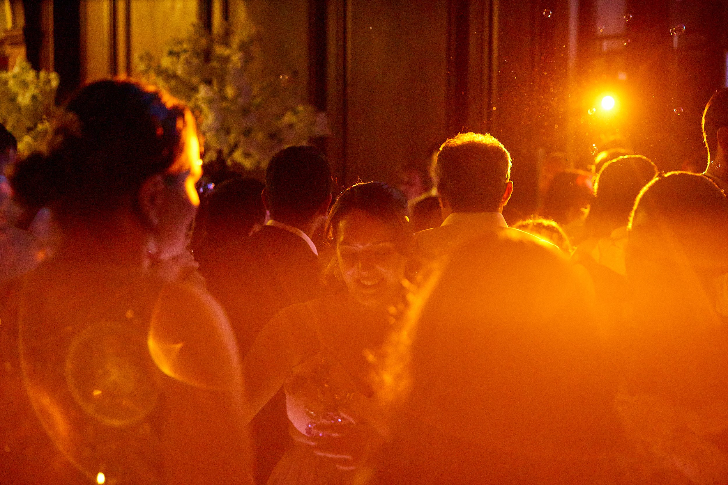 People socializing at an outdoor evening event, illuminated by warm orange and yellow lighting.