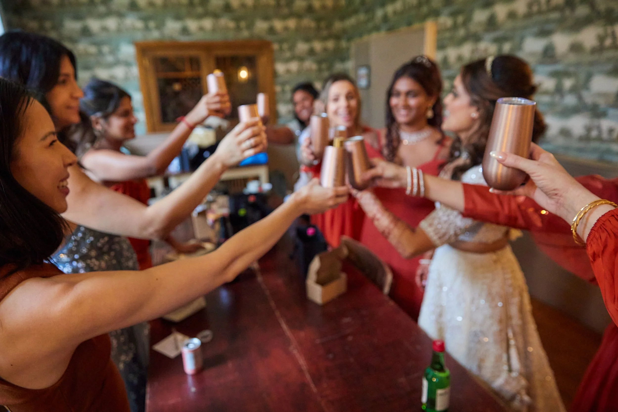 A group of women celebrating and raising copper-colored cups together in a festive setting, some dressed in traditional attire.