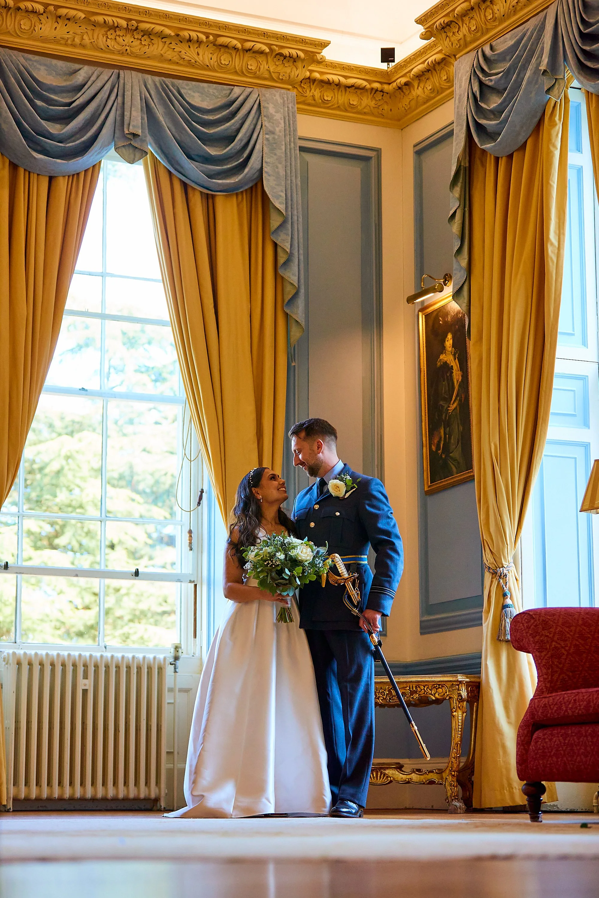A bride and groom share a moment inside a grand, elegant room with large windows and ornate curtains. The bride holds a bouquet and wears a white wedding dress, while the groom is dressed in a blue military uniform with a sword. They are gazing at ea