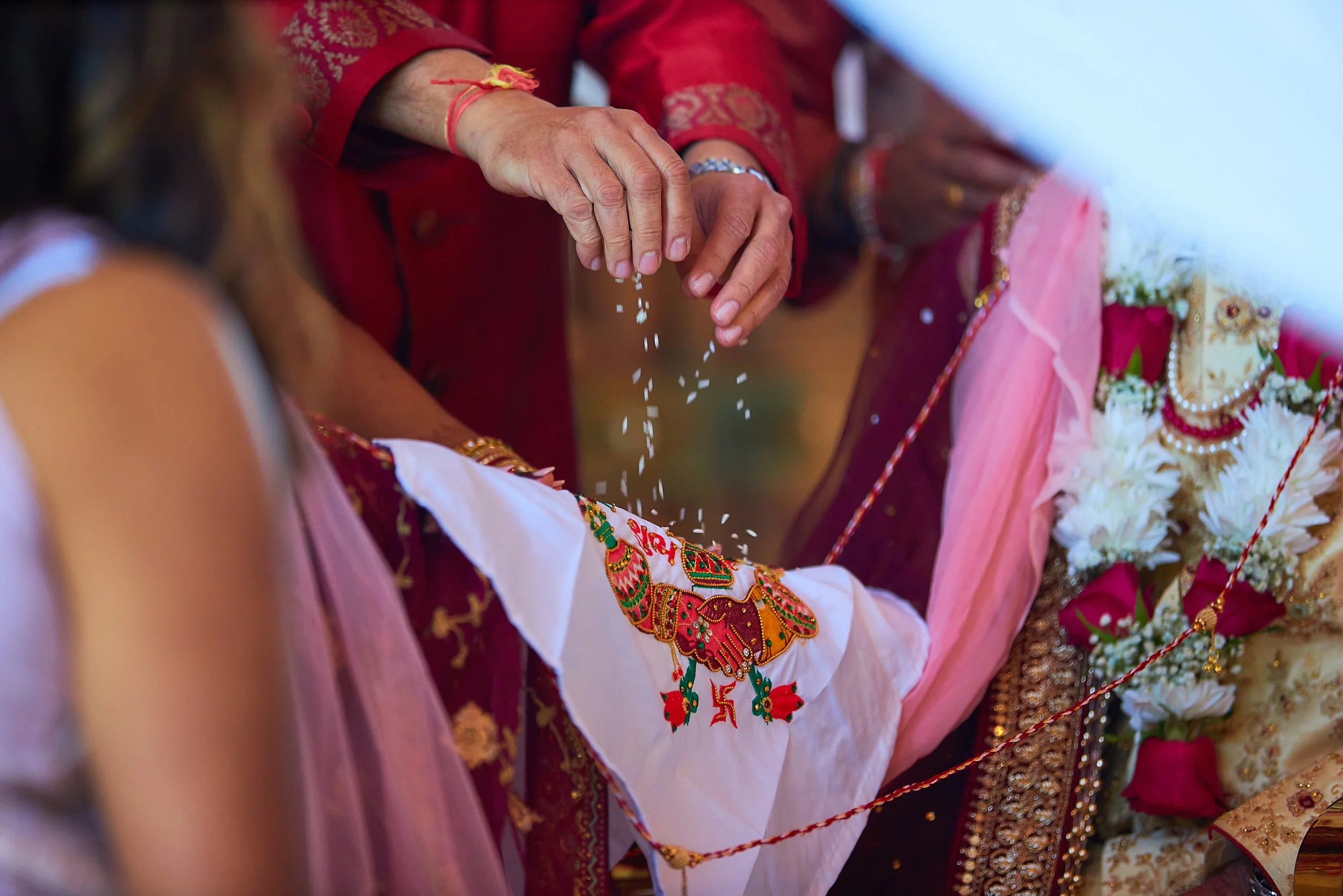 An elderly woman dressed in traditional red attire is sprinkling rice during a religious or cultural ceremony, with other women in colorful clothing and floral decorations in the background.