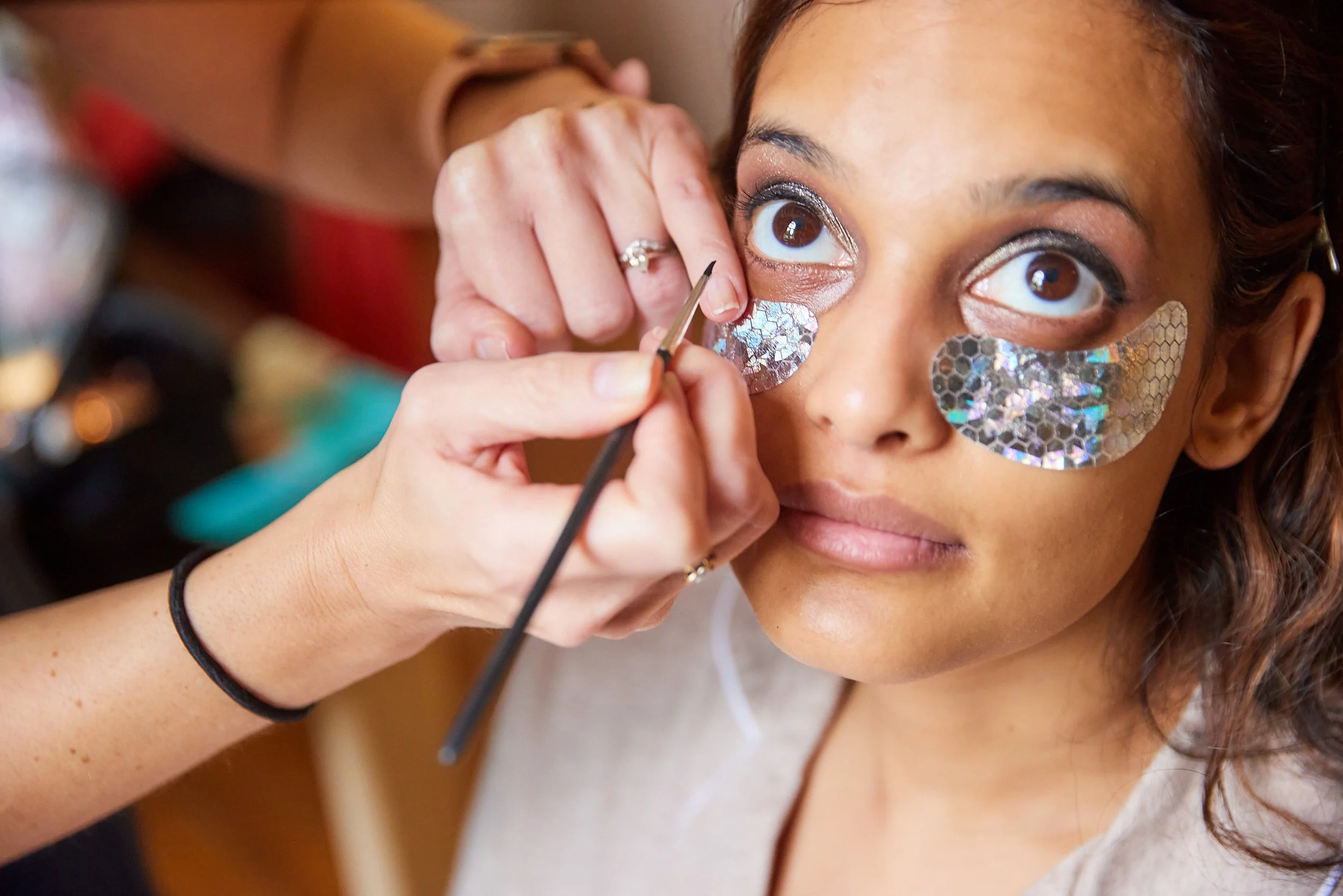 Woman receives makeup application with holographic under-eye patches, while makeup artist applies eyeliner under her eye.