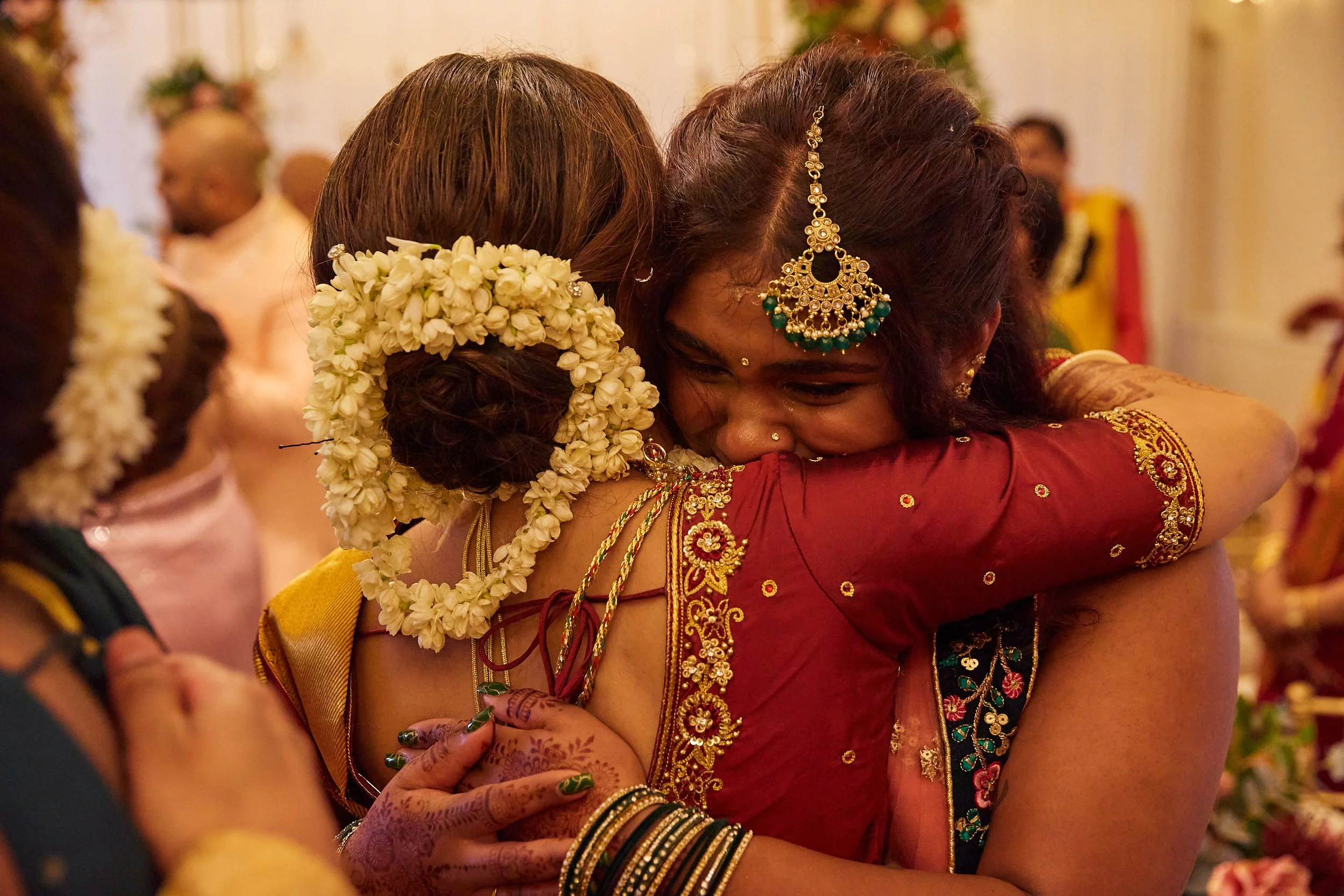 Two women embracing at a celebration, with one woman wearing traditional Indian attire and jewelry, and the other woman with a floral hair accessory.