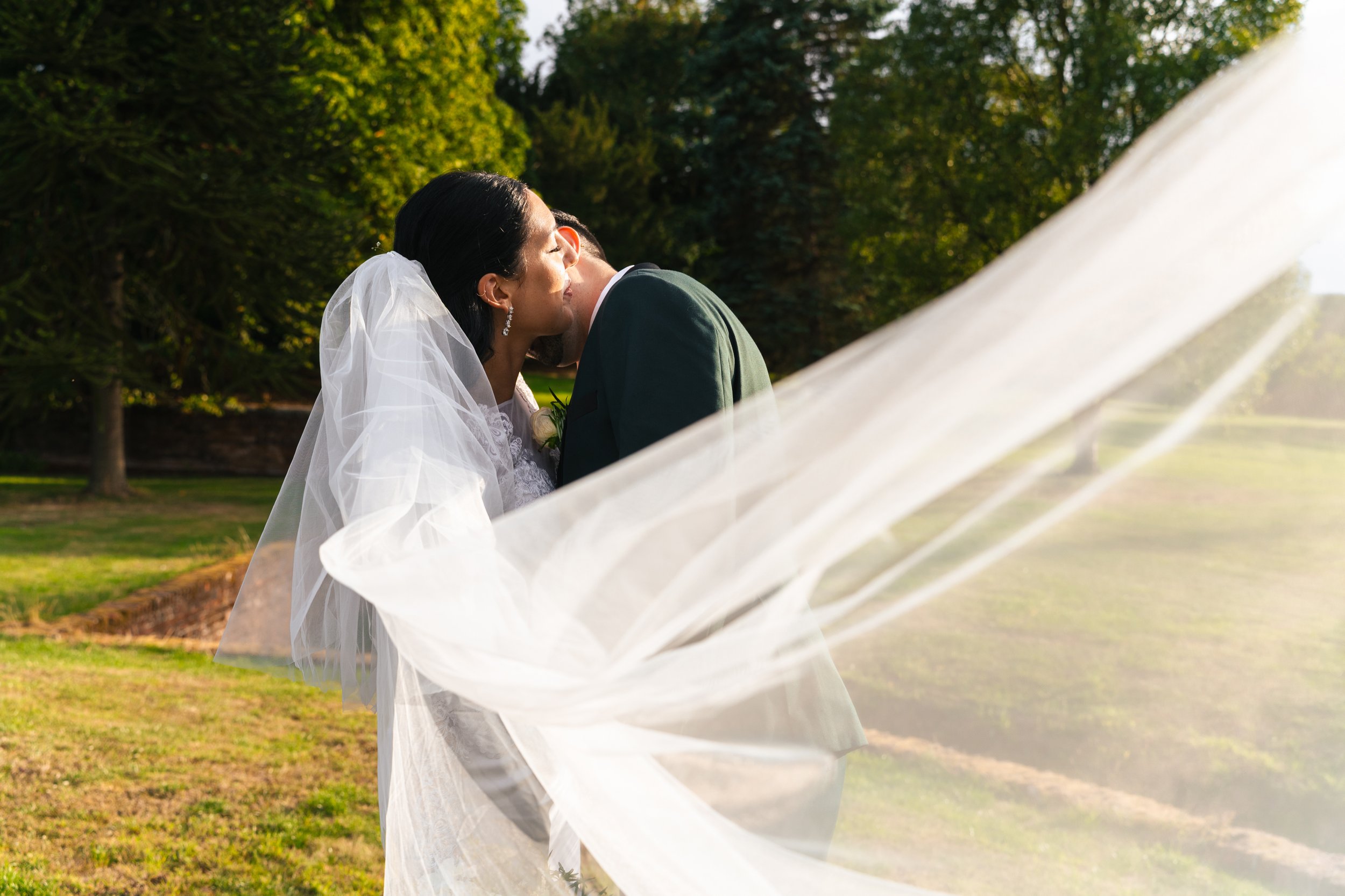 A bride and groom sharing a kiss outdoors on a sunny day, with the bride wearing a white wedding gown and veil, and the groom in a dark suit, surrounded by green trees and grass.