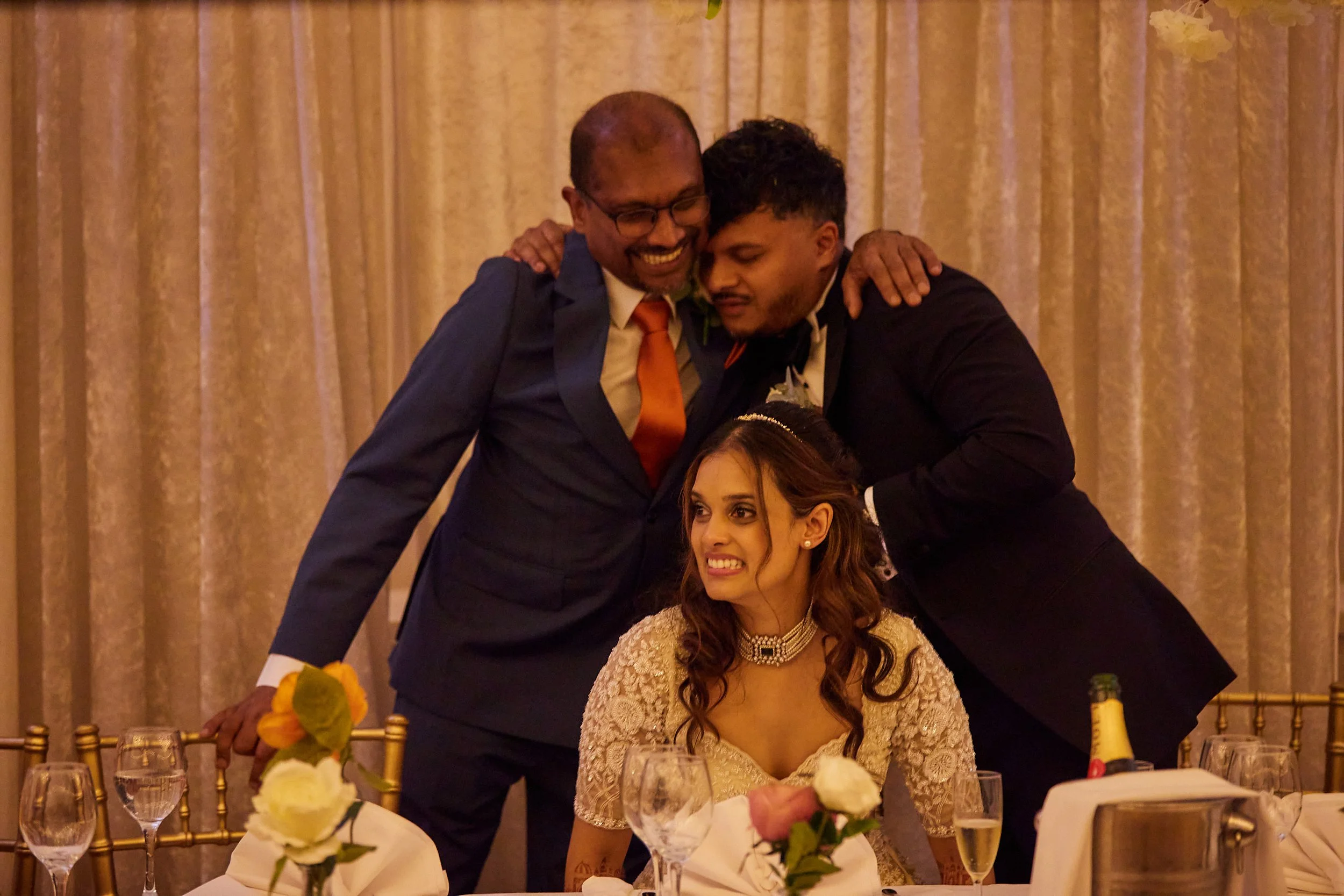 A woman in an elegant dress and choker necklace is sitting at a table, smiling and looking slightly to the side. Two men in suits are standing behind her, leaning over and hugging her shoulders, with one smiling broadly and the other with a more mute