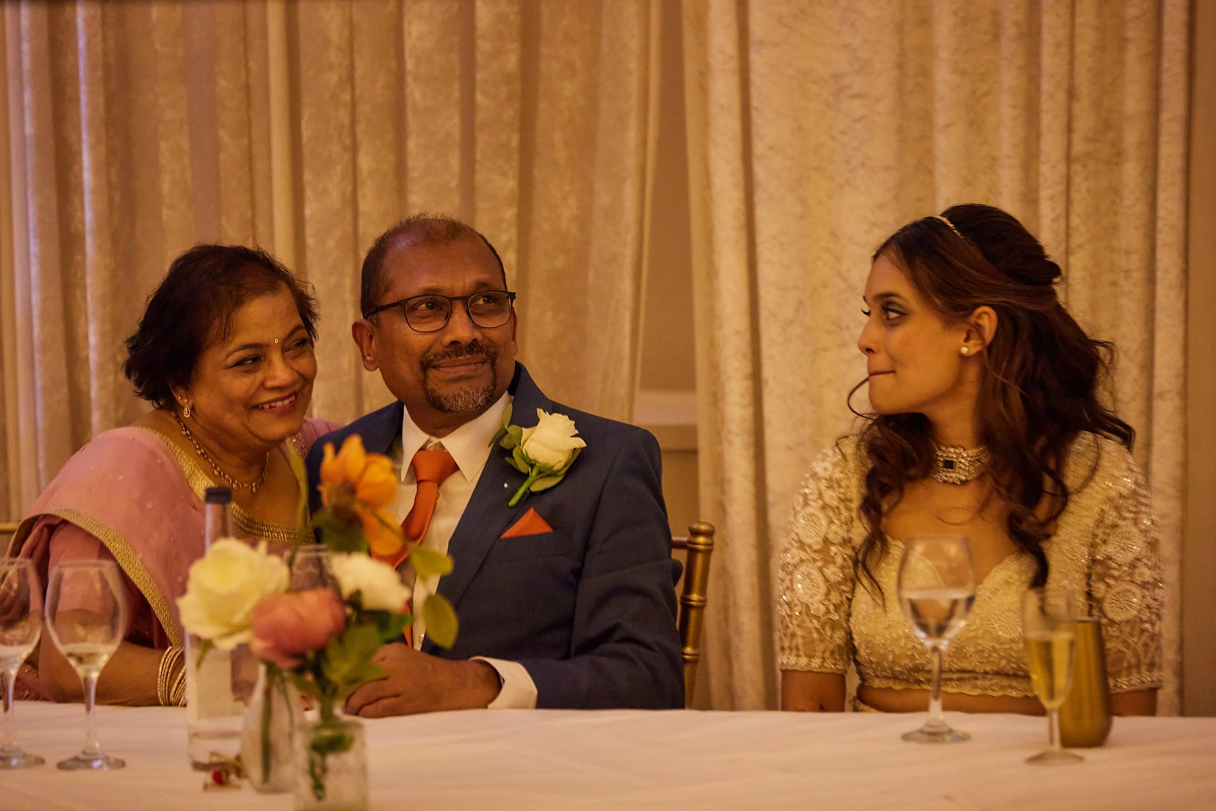 Three people sitting at a table during a formal event or celebration. The woman on the left is smiling, wearing a pink traditional dress and jewelry. The man in the middle is wearing a navy blue suit with an orange tie and glasses, looking to his rig