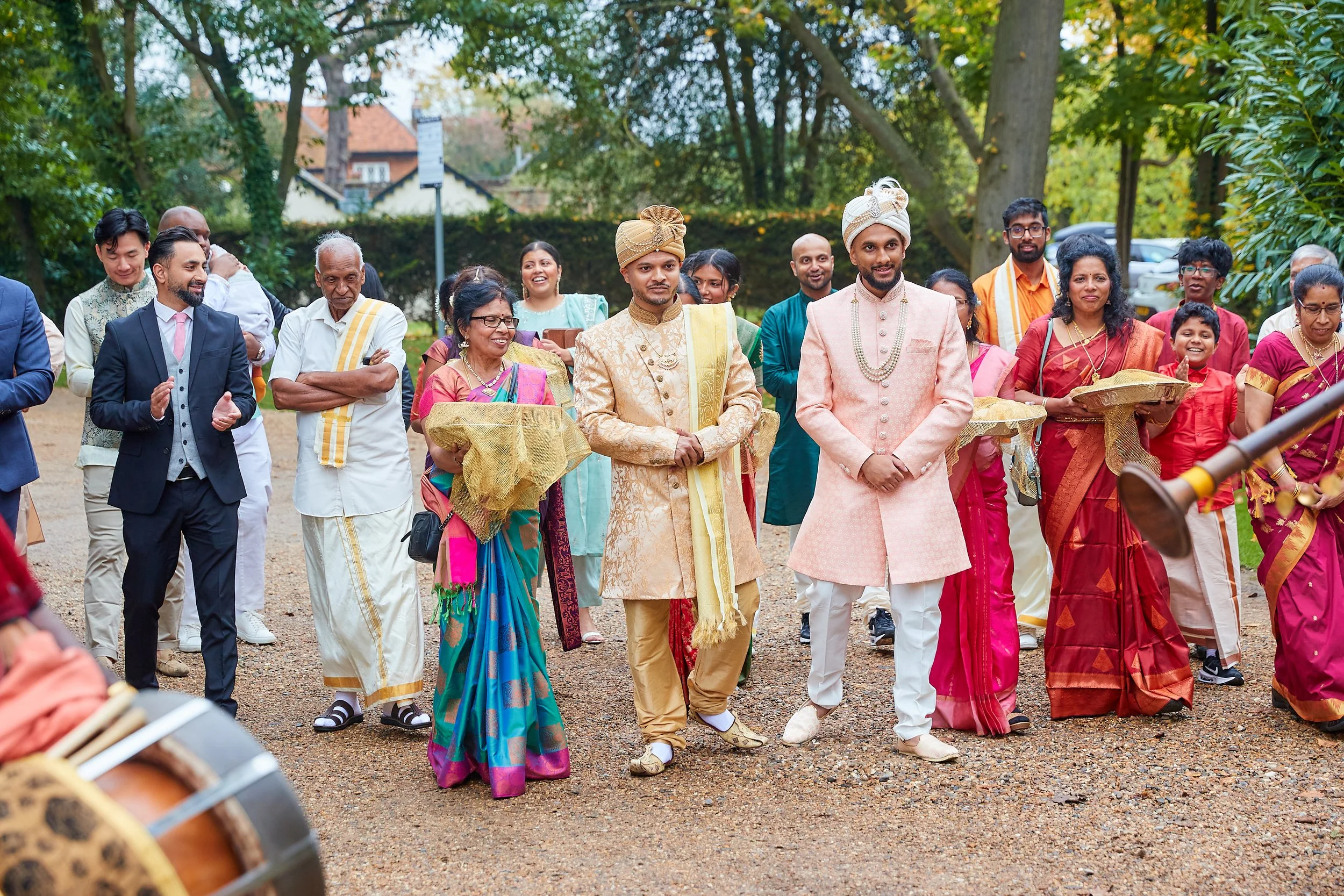 Group of people celebrating in traditional Indian attire, walking outdoors on a gravel path with trees and houses in the background.