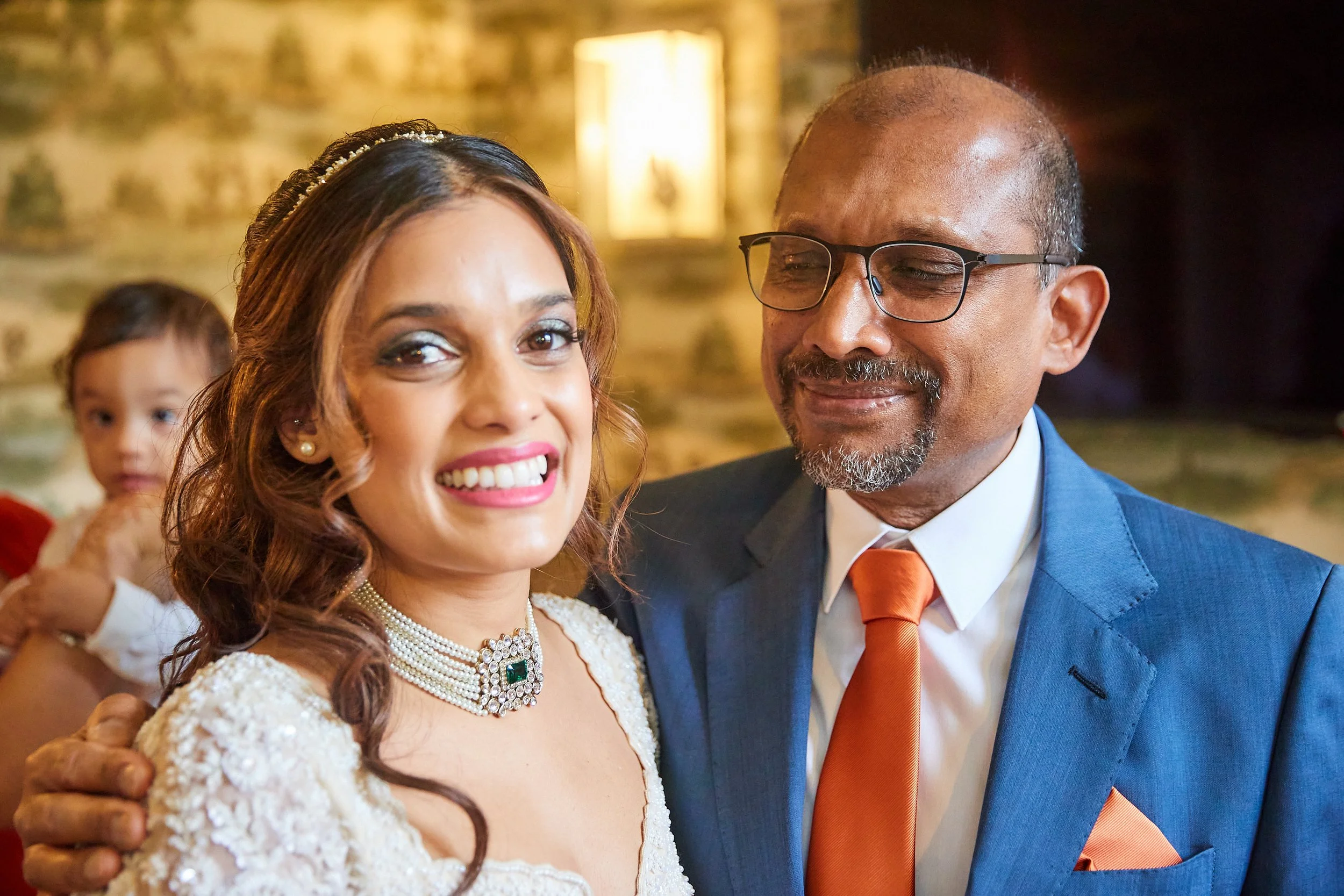Woman dressed in wedding gown standing next to a man in a blue suit with orange tie, smiling with a young girl in background.