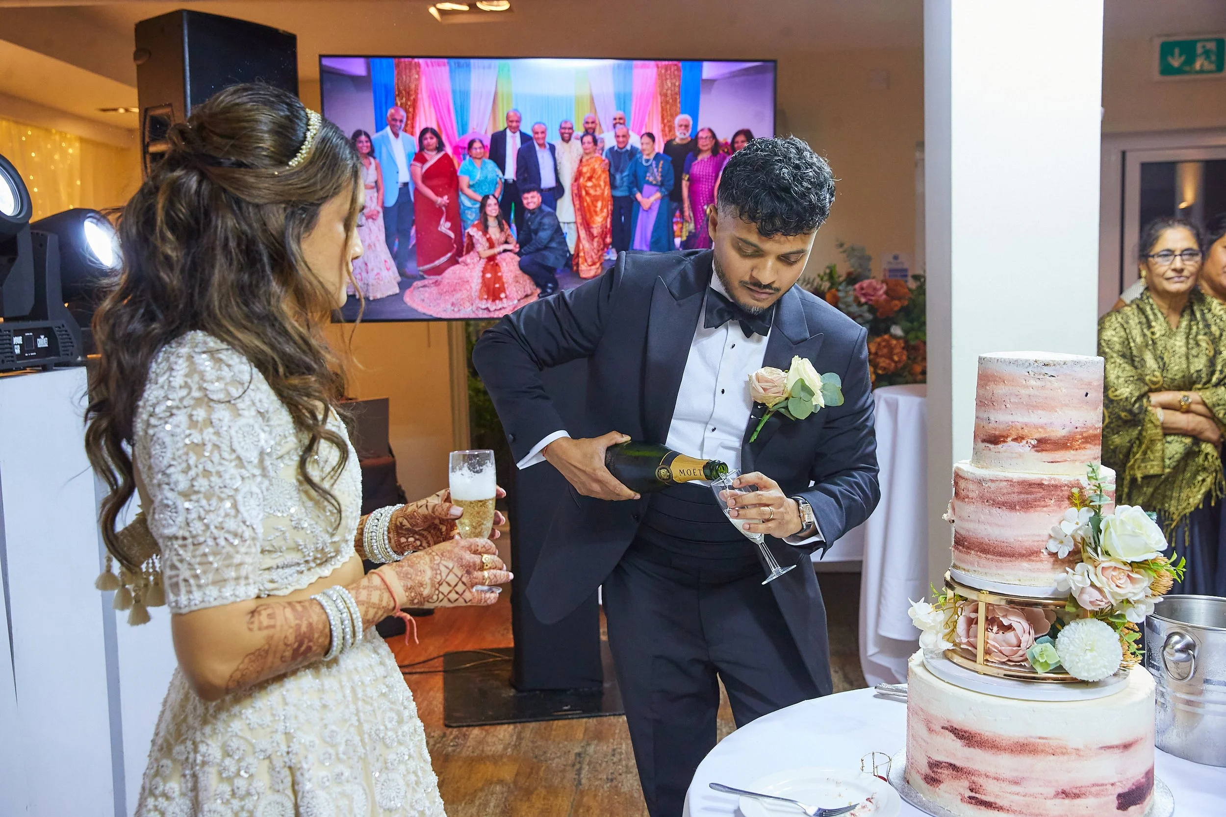 A man dressed in a black tuxedo pours champagne into a glass at a wedding celebration. A woman in a cream-colored dress with lace details and henna on her hands holds a flute of champagne. There is a large wedding cake decorated with flowers on the t