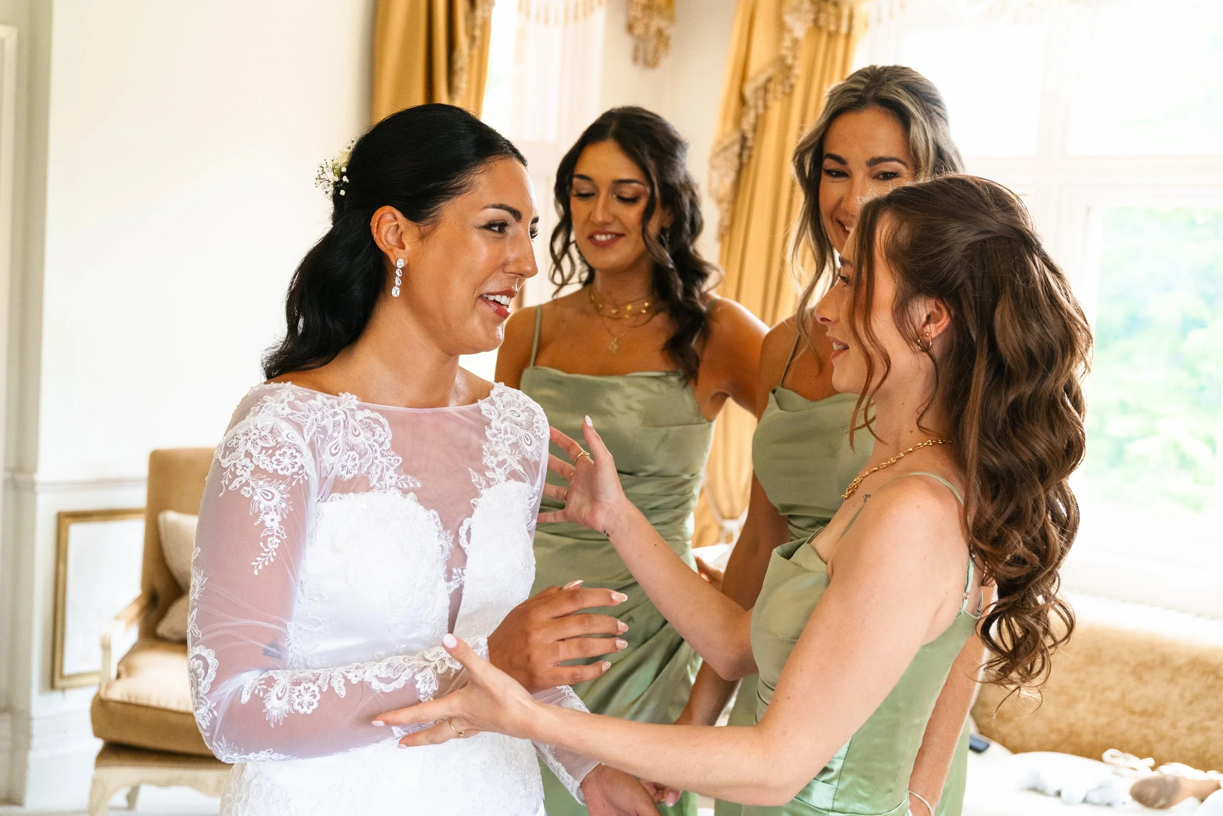 A bride in a white wedding dress surrounded by four women in green bridesmaid dresses, inside a bright room with gold curtains.