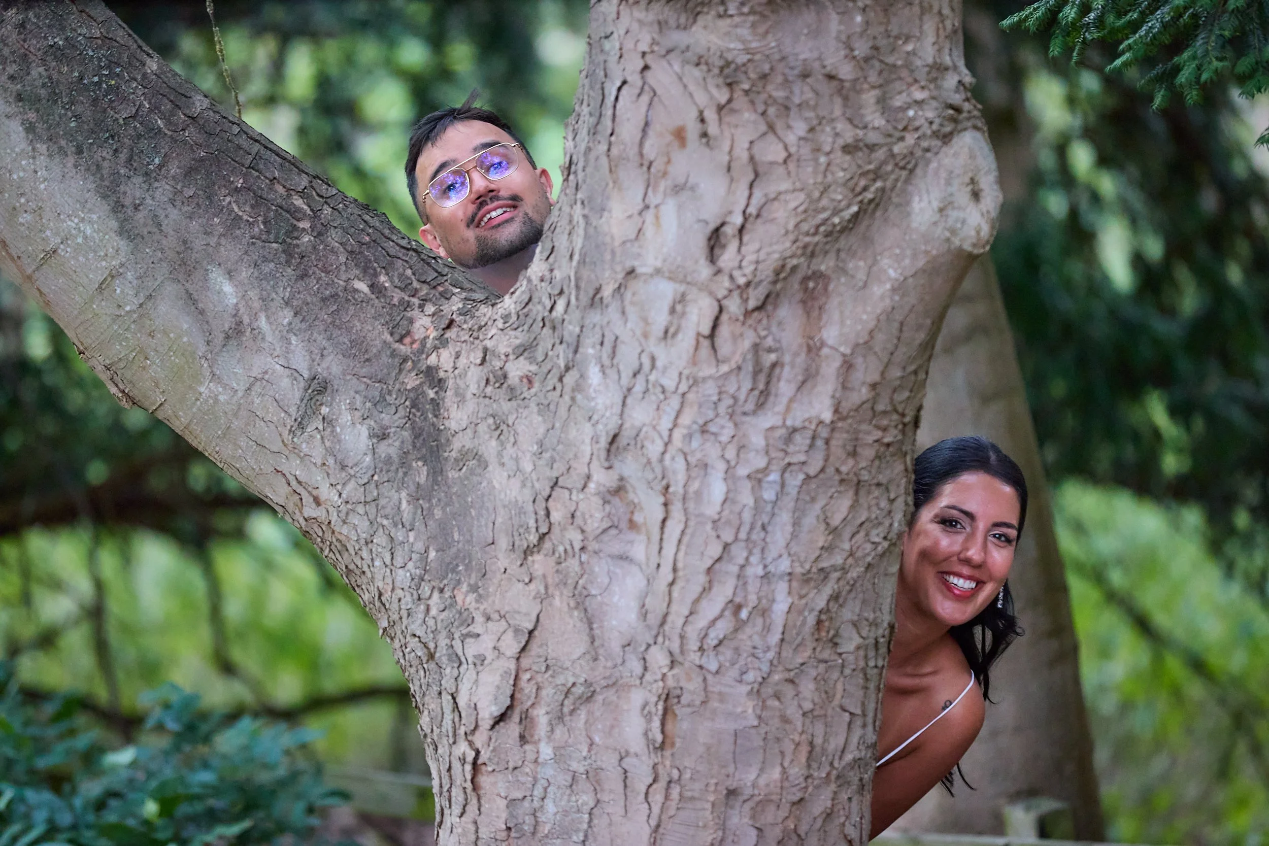 A man and woman peeking out from behind a large tree trunk in a green forest
