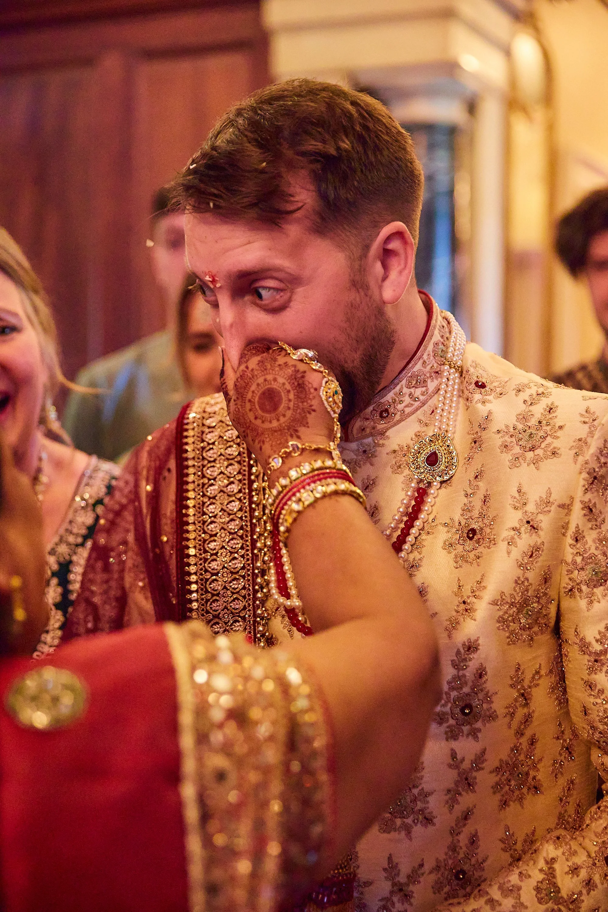 A man in traditional Indian attire at a wedding, covering his mouth with his hand, surrounded by women dressed in colorful Indian clothing, with intricate jewelry and henna designs on their hands.