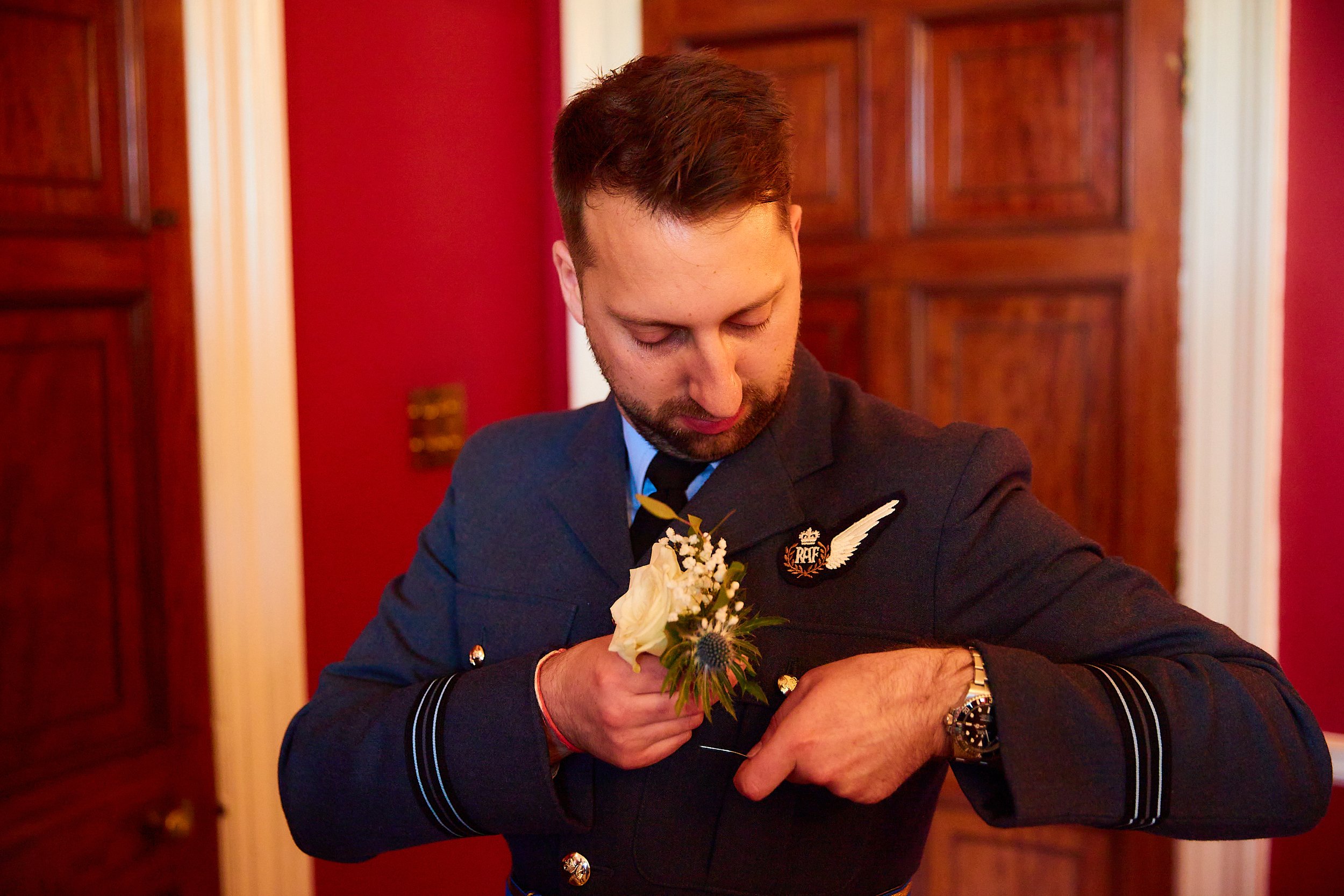 A man in a police uniform with a badge and wings pin, holding a white flower boutonniere while adjusting his sleeve, standing in a room with red walls and wooden paneling.