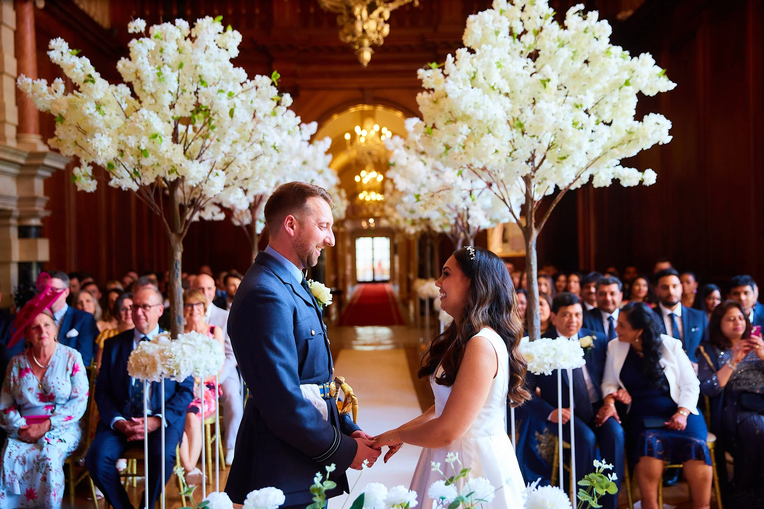 A bride and groom holding hands, smiling at each other during their wedding ceremony in a decorated church with white cherry blossom trees and a seated audience.