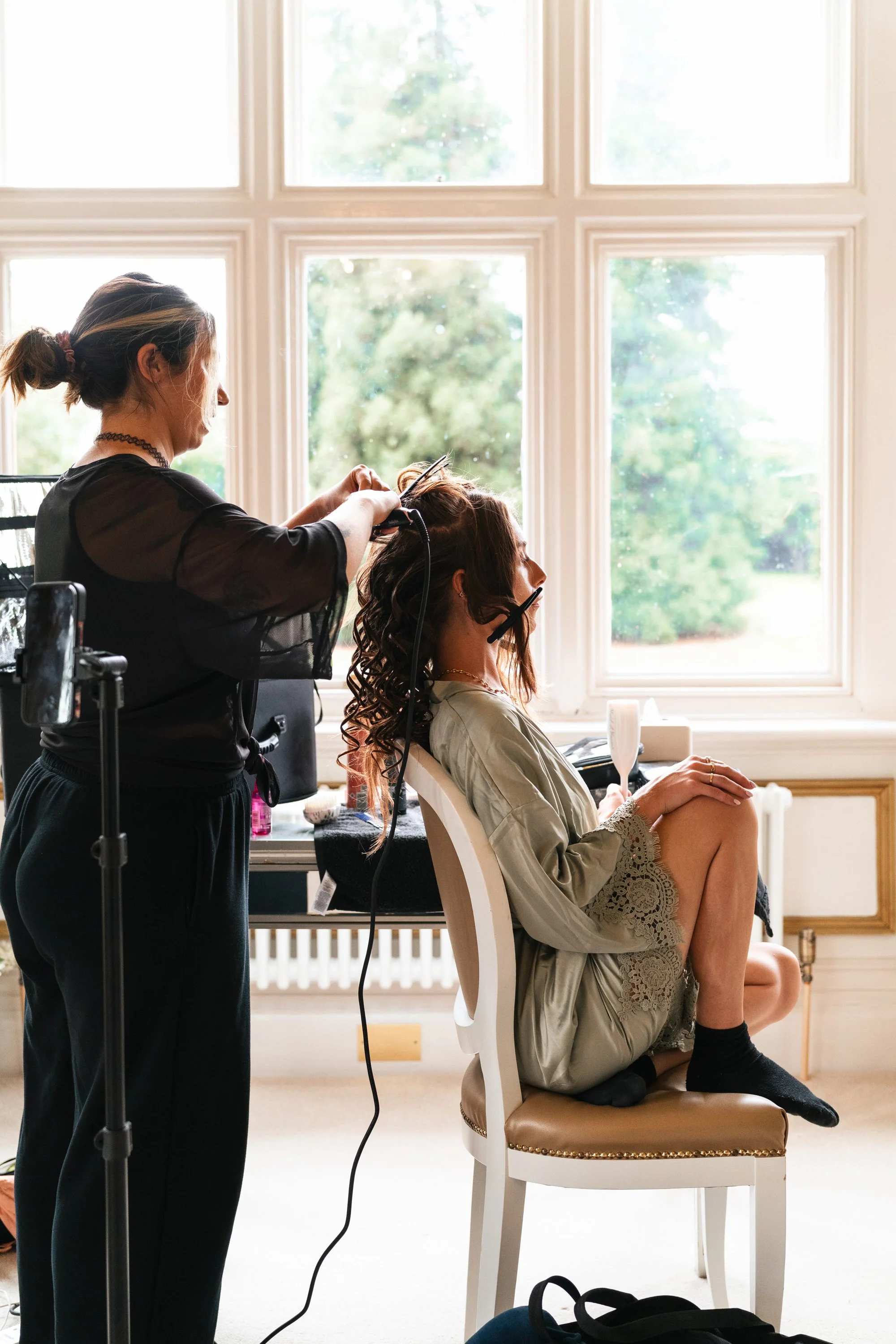 A woman getting her hair styled by a hairstylist in a bright room with large windows showing trees outside.