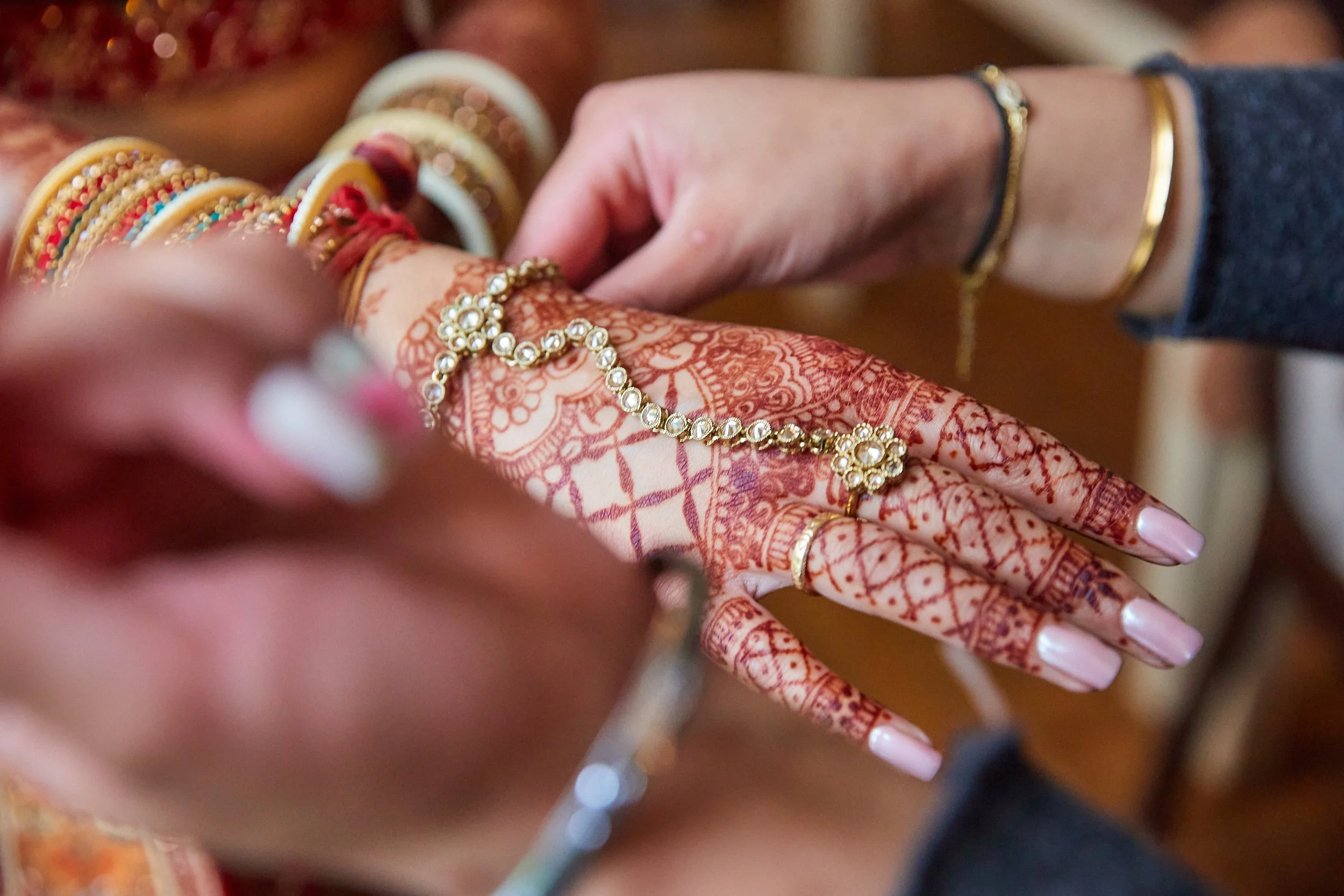 A person applying intricate henna designs to a woman's hand, who is wearing gold jewelry and colorful bangles, during a traditional celebration or wedding.