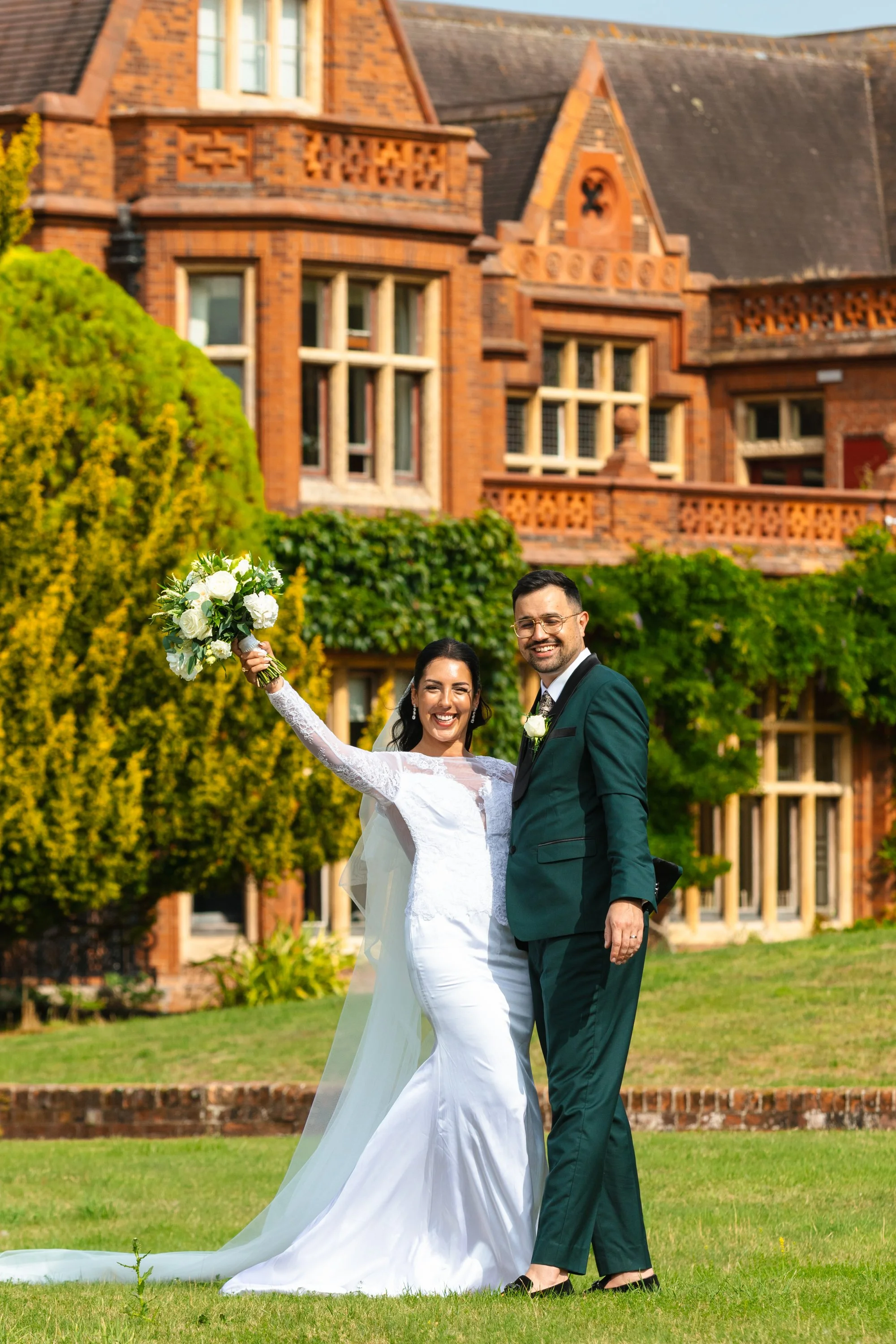 A newlywed couple stands on a lawn in front of a large red brick house. The bride is holding a bouquet of white flowers and has her arm raised, smiling. The groom is smiling, wearing a green suit with a white boutonniere. The scene is bright and chee