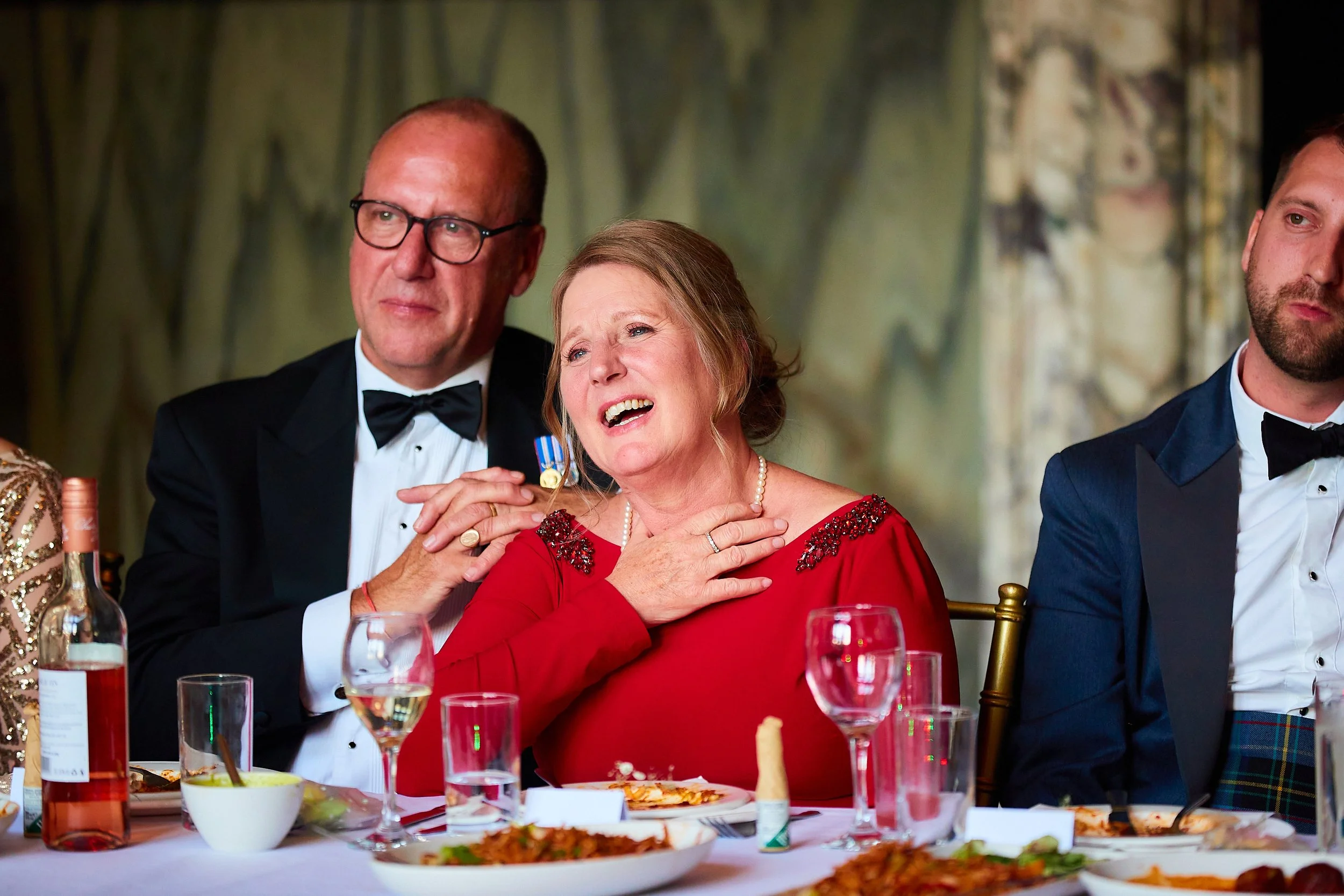 A woman in a red dress is sitting at a table, appearing emotional, with her hand on her chest. Two men in tuxedos are sitting next to her, one touching her shoulder. There are glasses and plates on the table, suggesting a formal dinner event.