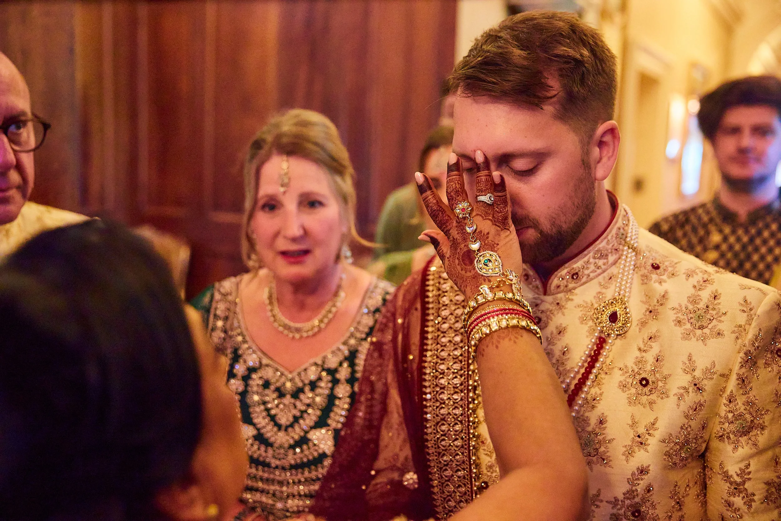 A traditional Indian wedding ceremony with a woman applying henna to a groom's face. The groom is dressed in an ornate cream-colored sherwani with gold embroidery, and is wearing jewelry. The woman has henna on her hands, adorned with rings, bangles,
