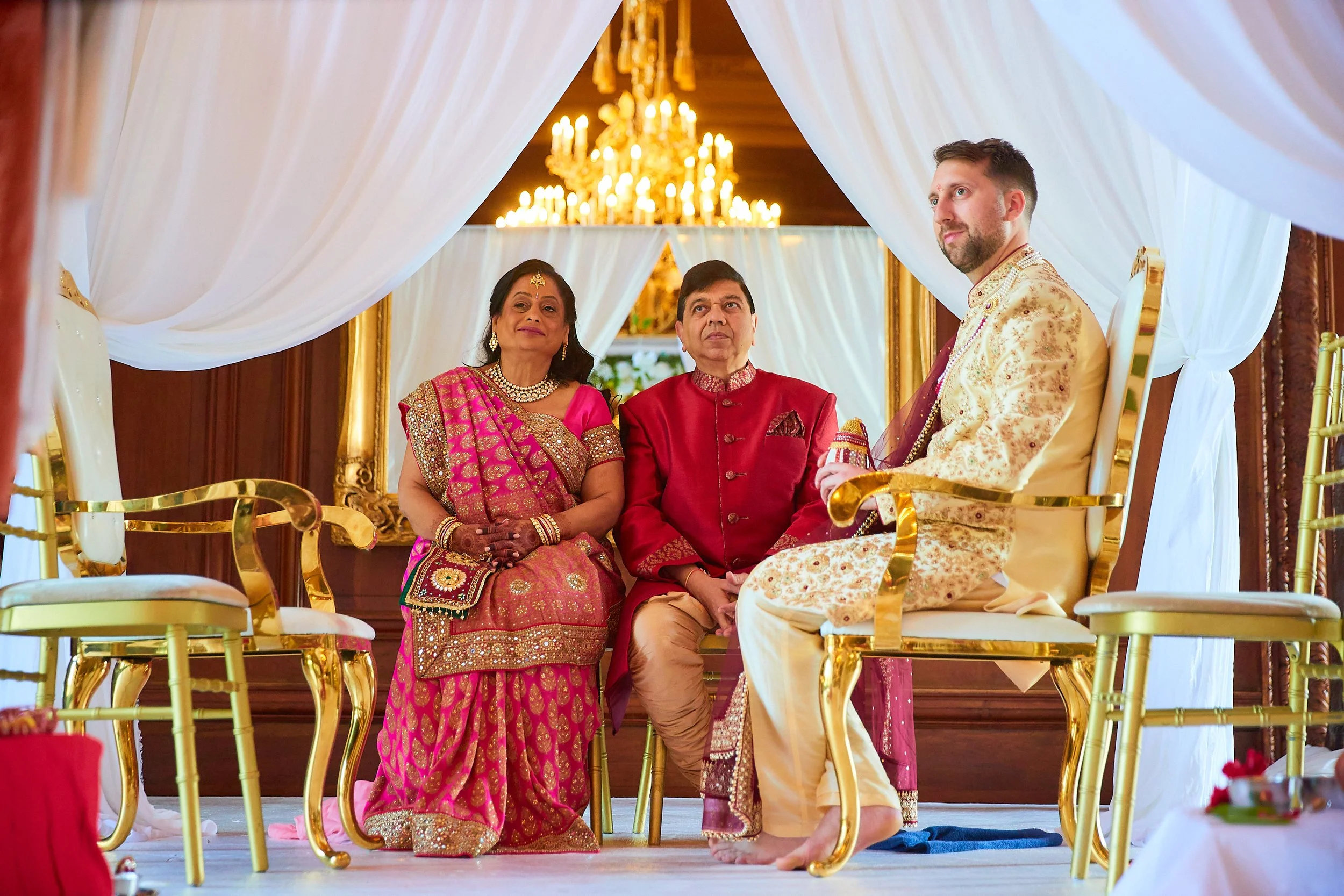 Indian wedding ceremony with couple dressed in traditional wedding attire sitting on platform under white drapes, surrounded by family members, with chandelier overhead.