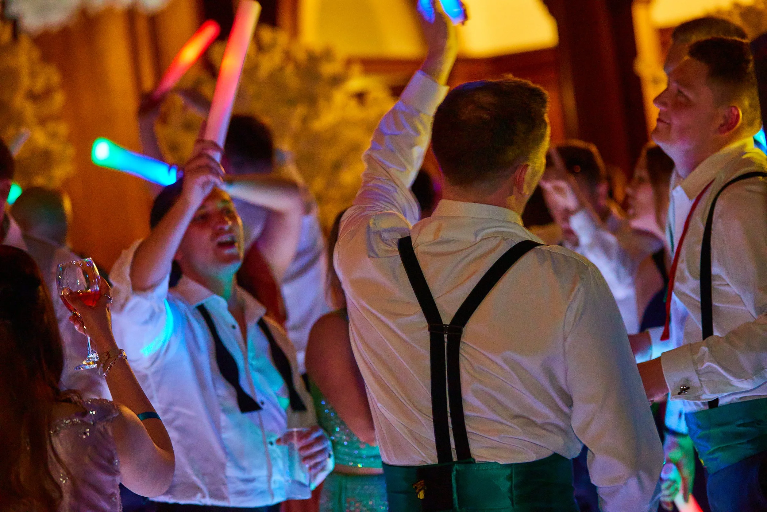 People celebrating at a party with colorful glow sticks and drinks, dressed in formal or semi-formal attire, in a lively indoor setting.