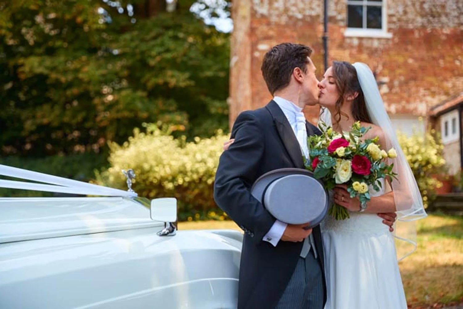A bride and groom kissing outside near a vintage white car, with the bride holding a bouquet of red, white, and yellow flowers, surrounded by greenery and autumn foliage.