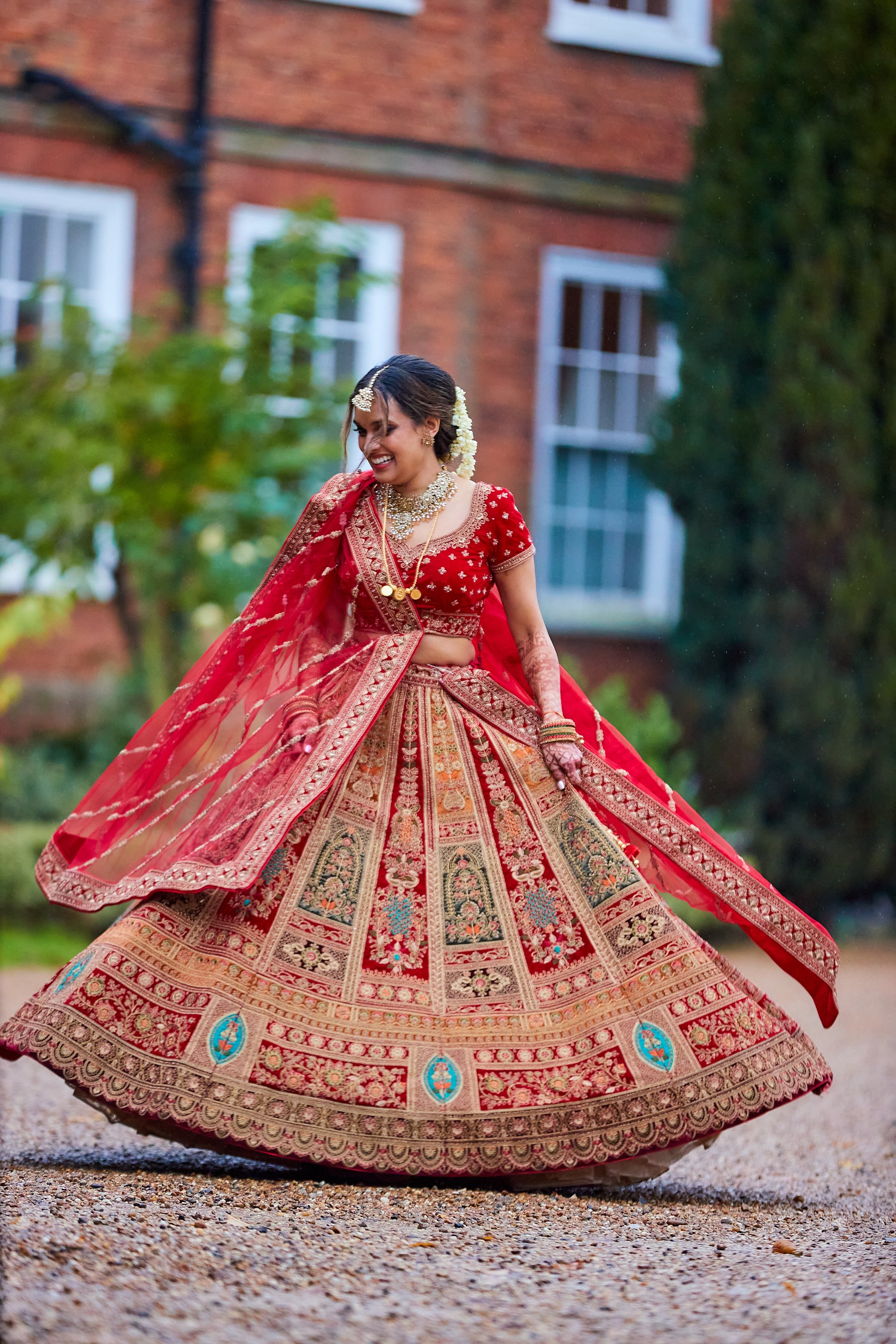 A woman in a vibrant red traditional Indian bridal outfit, twirling outdoors with a brick house and greenery in the background.