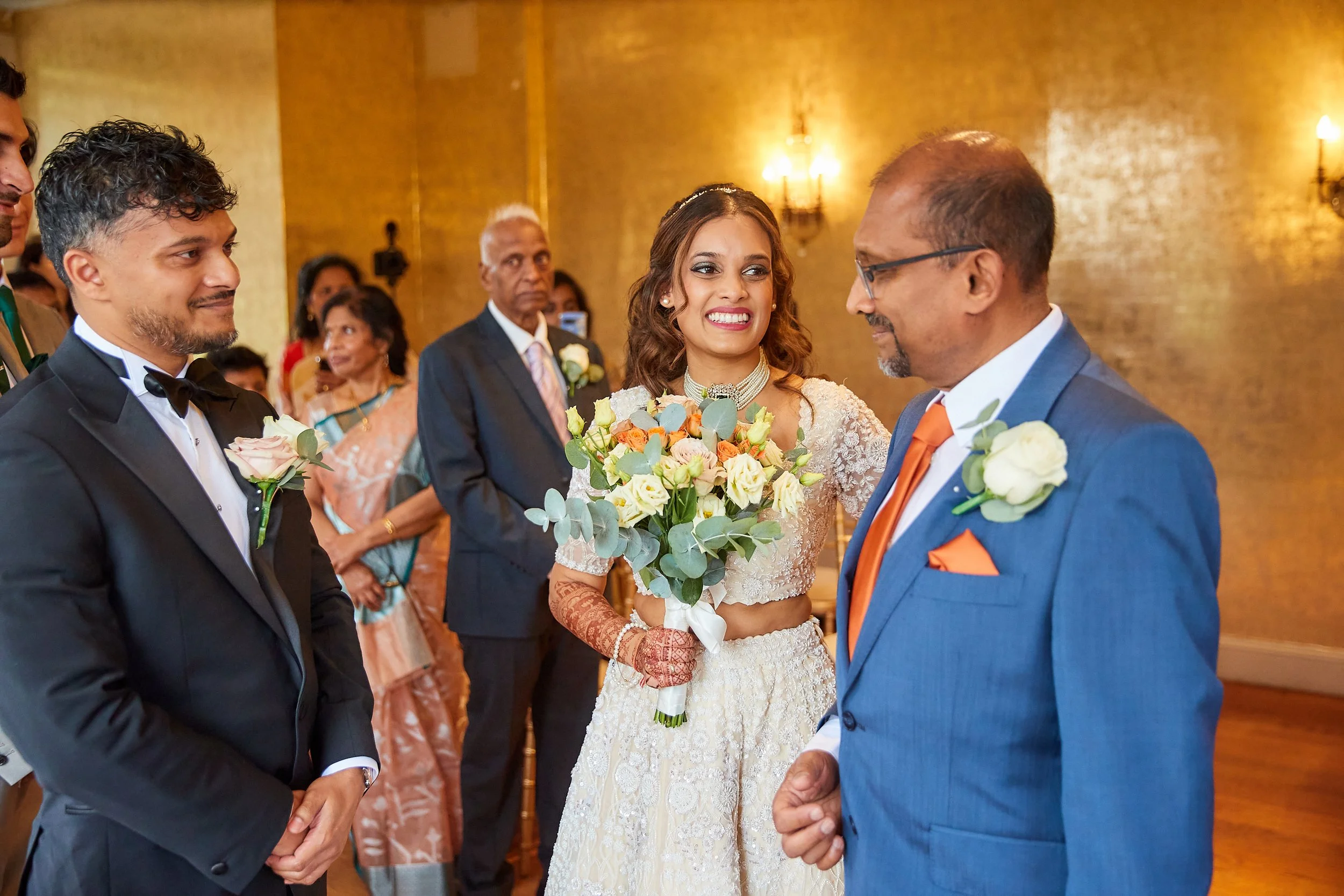A bride in a lace wedding dress holding a bouquet, smiling. She stands next to a man in a blue suit with an orange tie and white boutonniere. Two men in tuxedos are on the left, one smiling. In the background, guests are watching the ceremony, with a