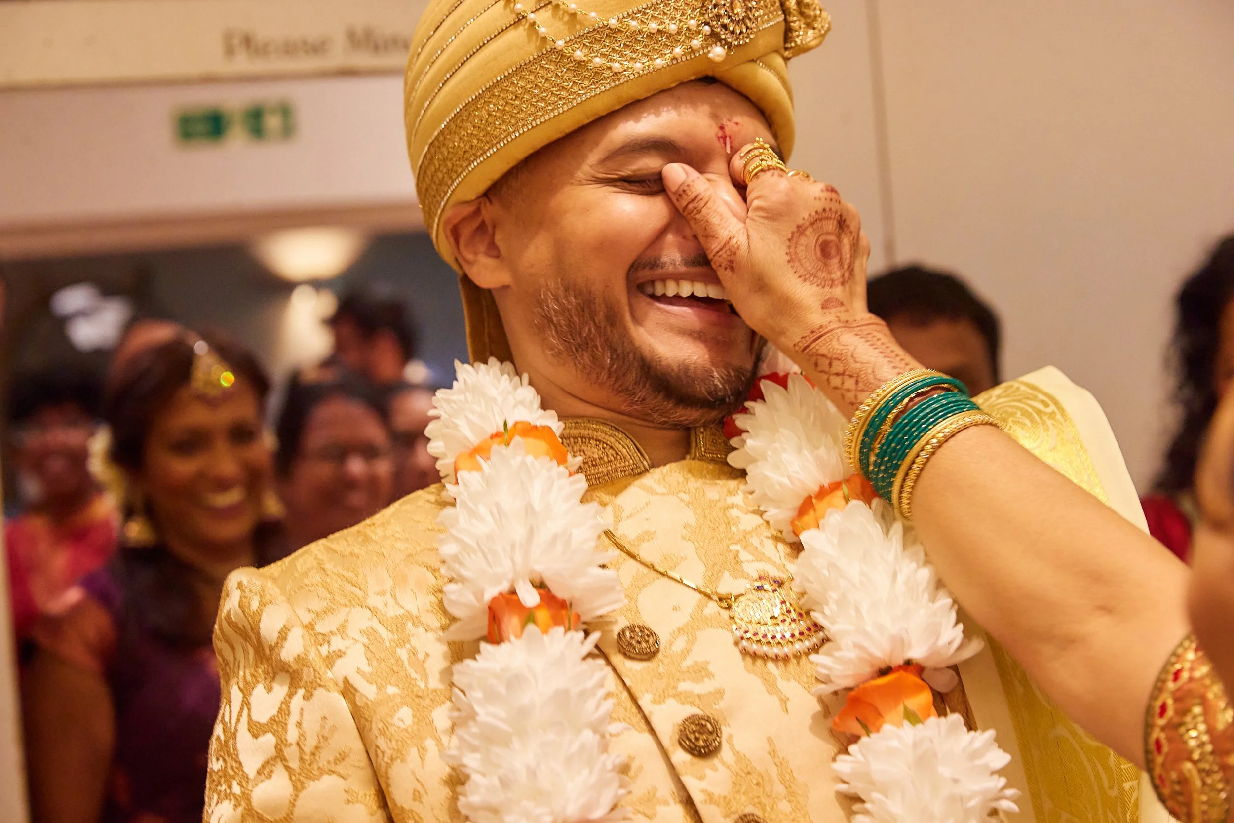 A joyful man dressed in traditional Indian wedding attire, wearing a gold turban, ornate gold sherwani, and flower garlands, is being touched on the nose by a woman whose hands are decorated with henna. The man is smiling with his eyes closed, and pe