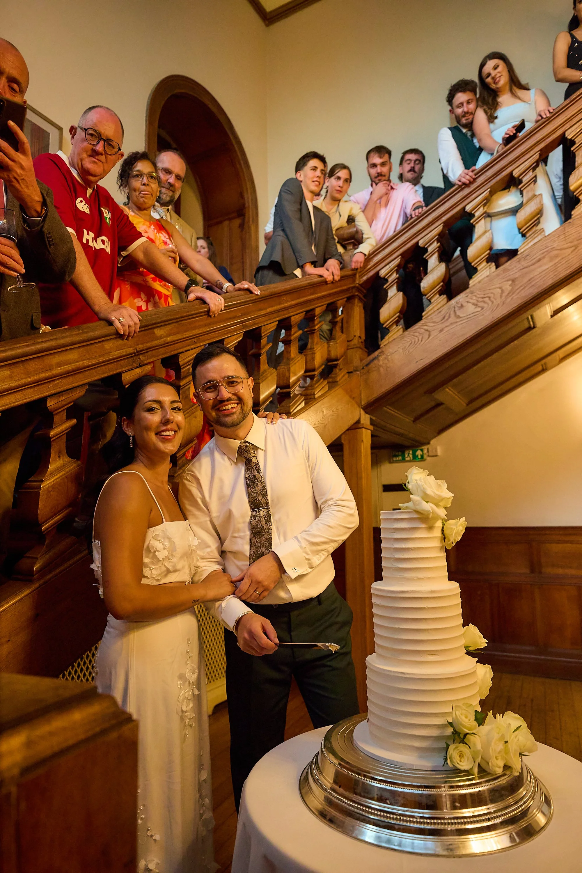 A newlywed couple cutting their wedding cake, with friends and family gathered on a wooden staircase behind them, in an elegant indoor setting.