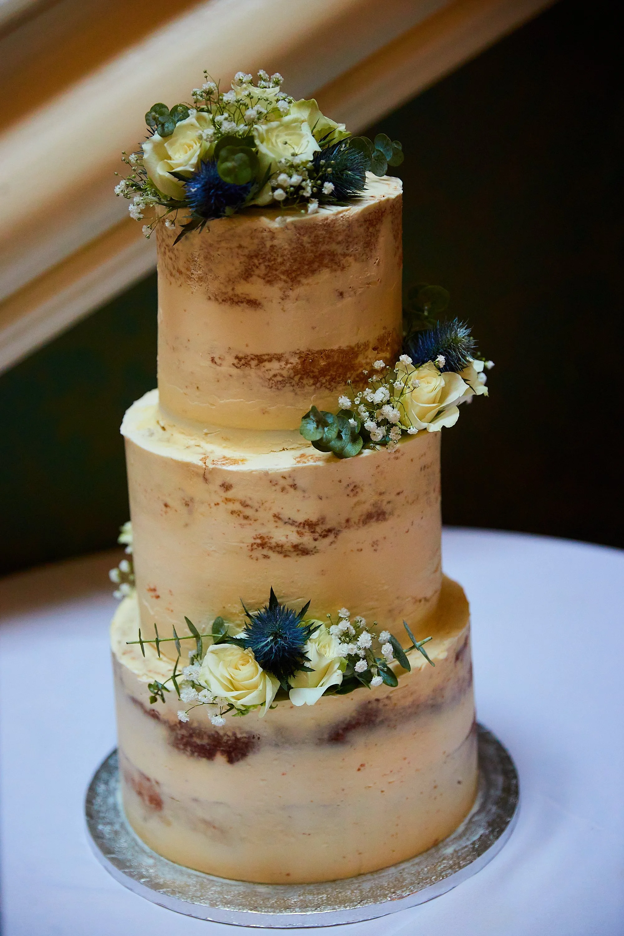 Three-tiered wedding cake decorated with white roses, small white flowers, green foliage, and blue thistle flowers, on a white tablecloth.
