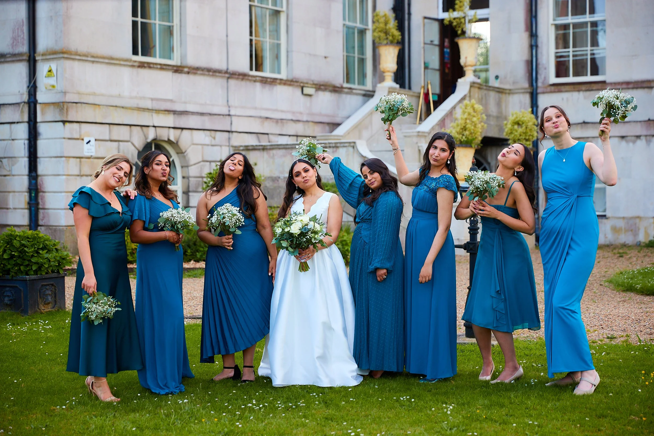 Group of eight women, including a bride in a white wedding dress, posing outdoors in front of a historic building, wearing matching blue dresses, holding bouquets, celebrating at a wedding.