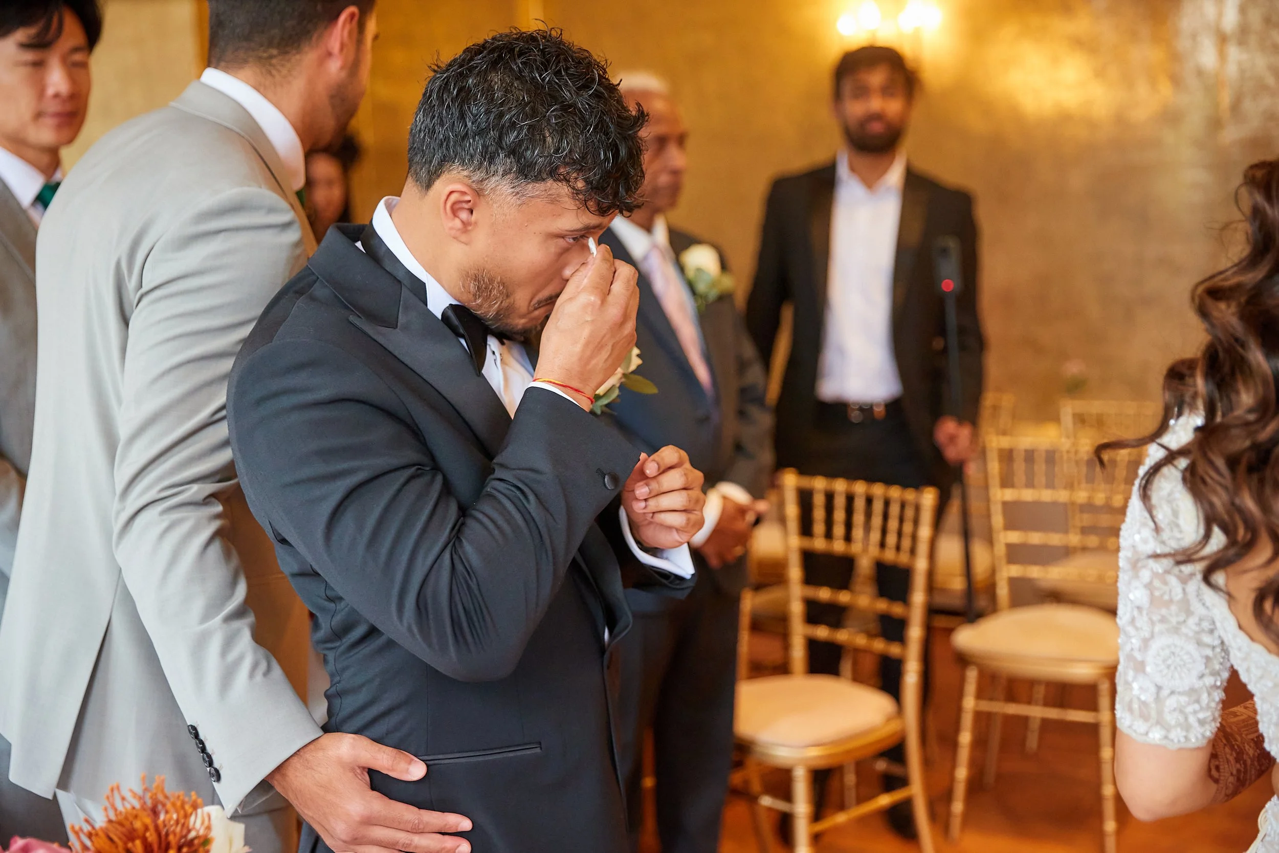 A man in a black tuxedo is crying and wiping his eyes while another man in a tan suit gently holds his back during a wedding ceremony.