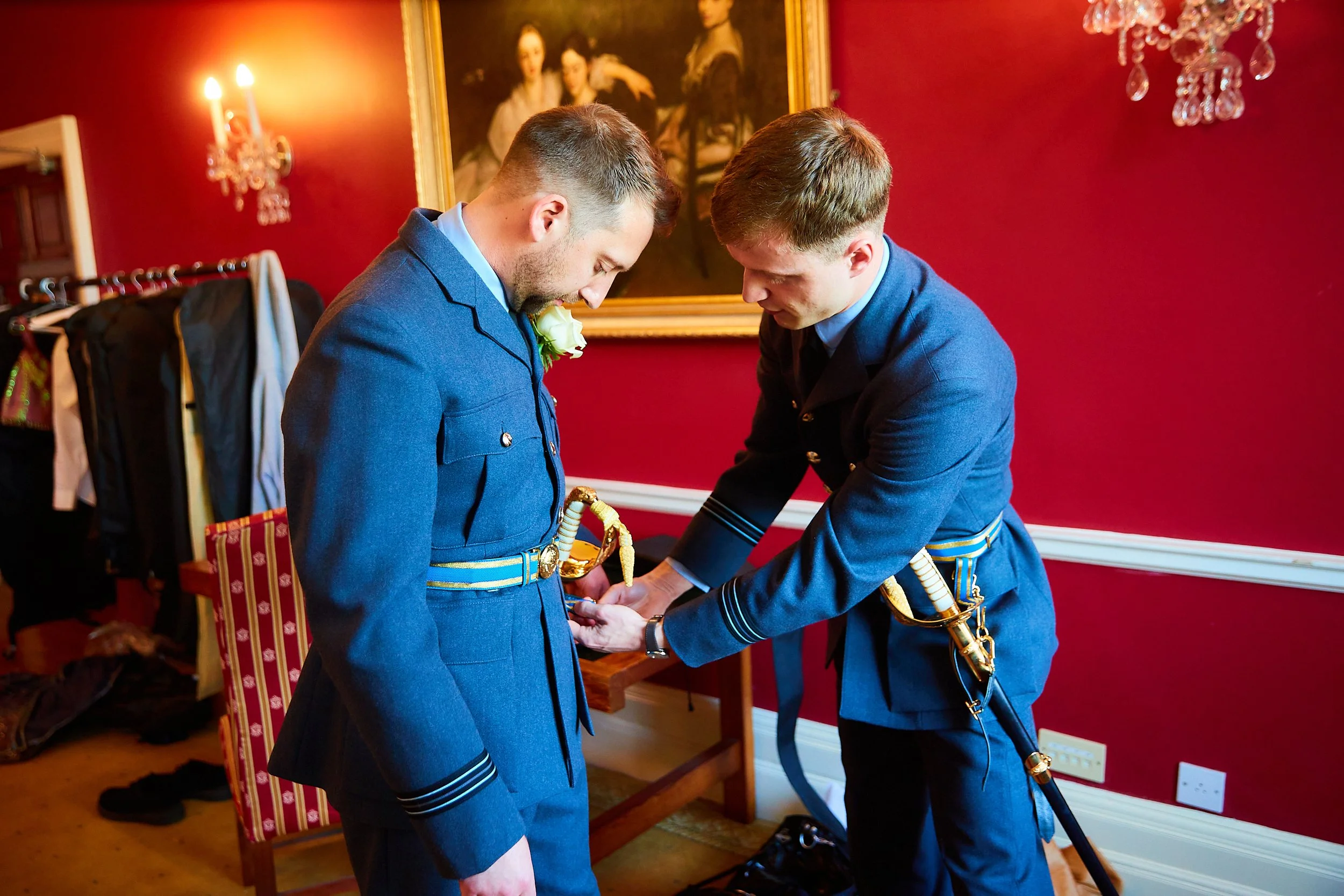Two men in Blue military uniforms, one with a sword, are standing in a room with red walls, and are involved in a ceremonial activity, possibly a decoration or presentation of a badge or medal.