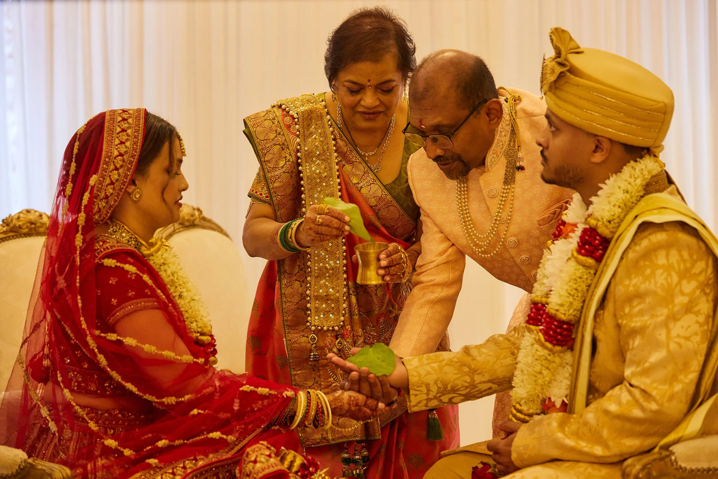 Indian wedding ceremony with bride and groom seated, engaging with family members, with gold and red traditional attire and floral garlands.