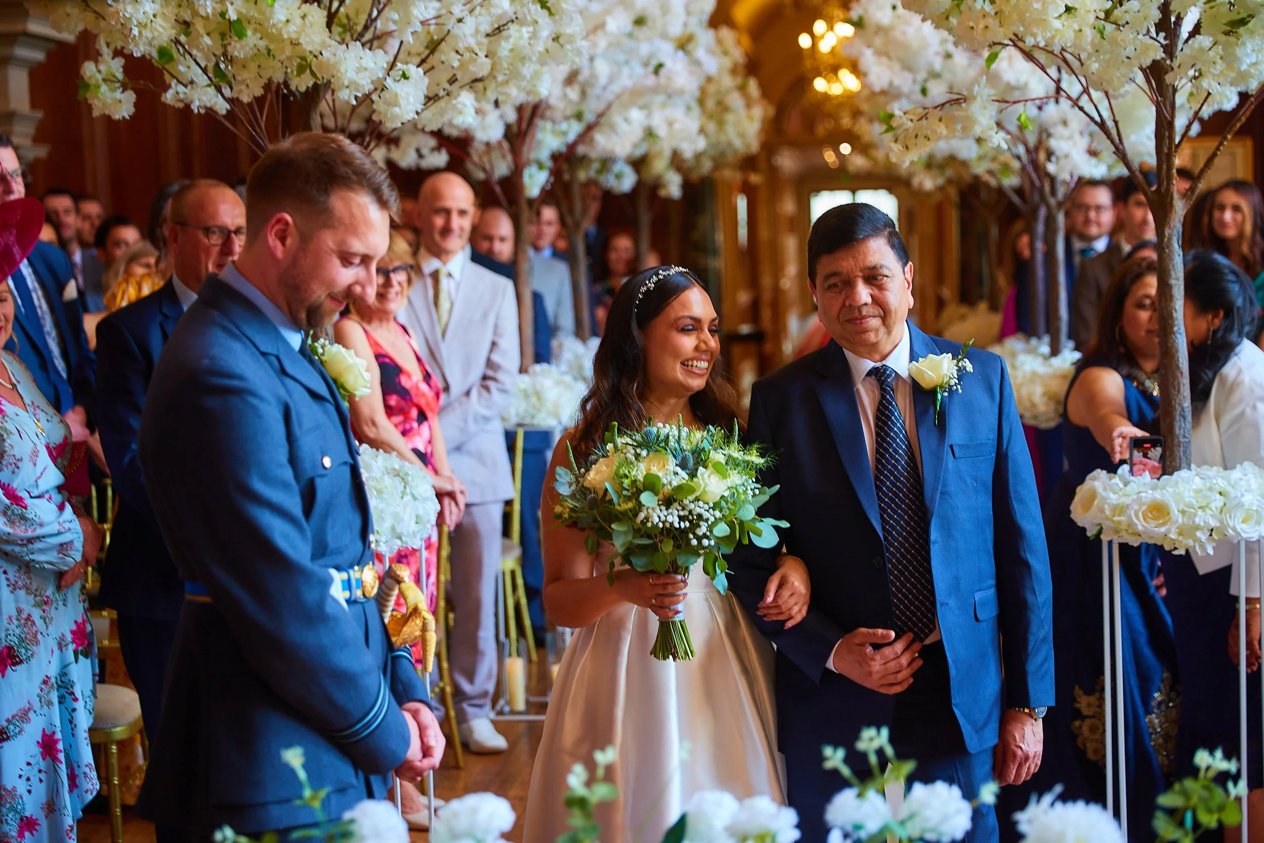 Bride being walked down the aisle by her father during a wedding ceremony, surrounded by guests and floral decorations.