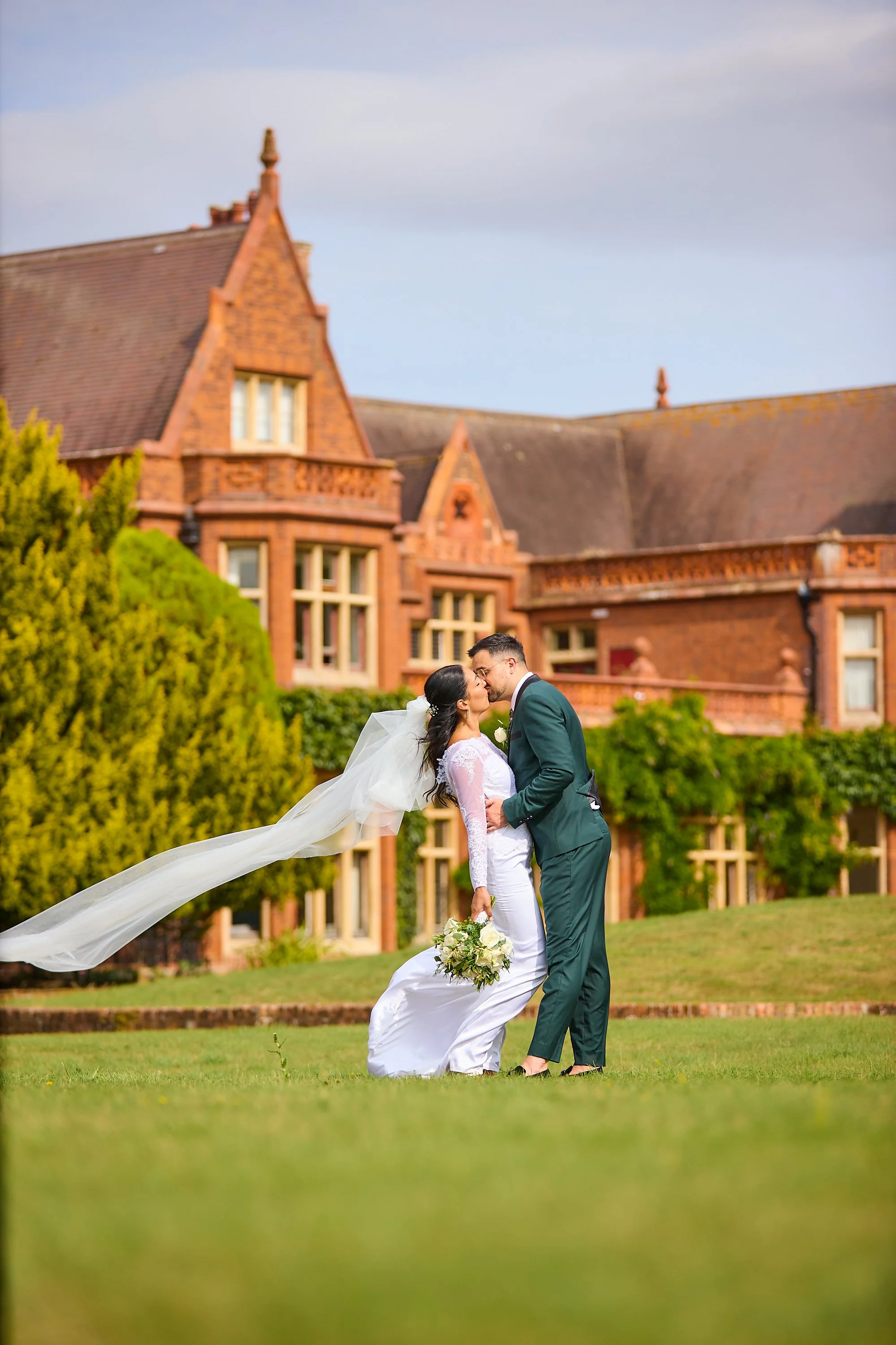Bride and groom kissing outdoors in front of a large brick house, groom wearing a green suit, bride in a white wedding dress holding a bouquet, bride's veil flowing in the wind, lush greenery around.