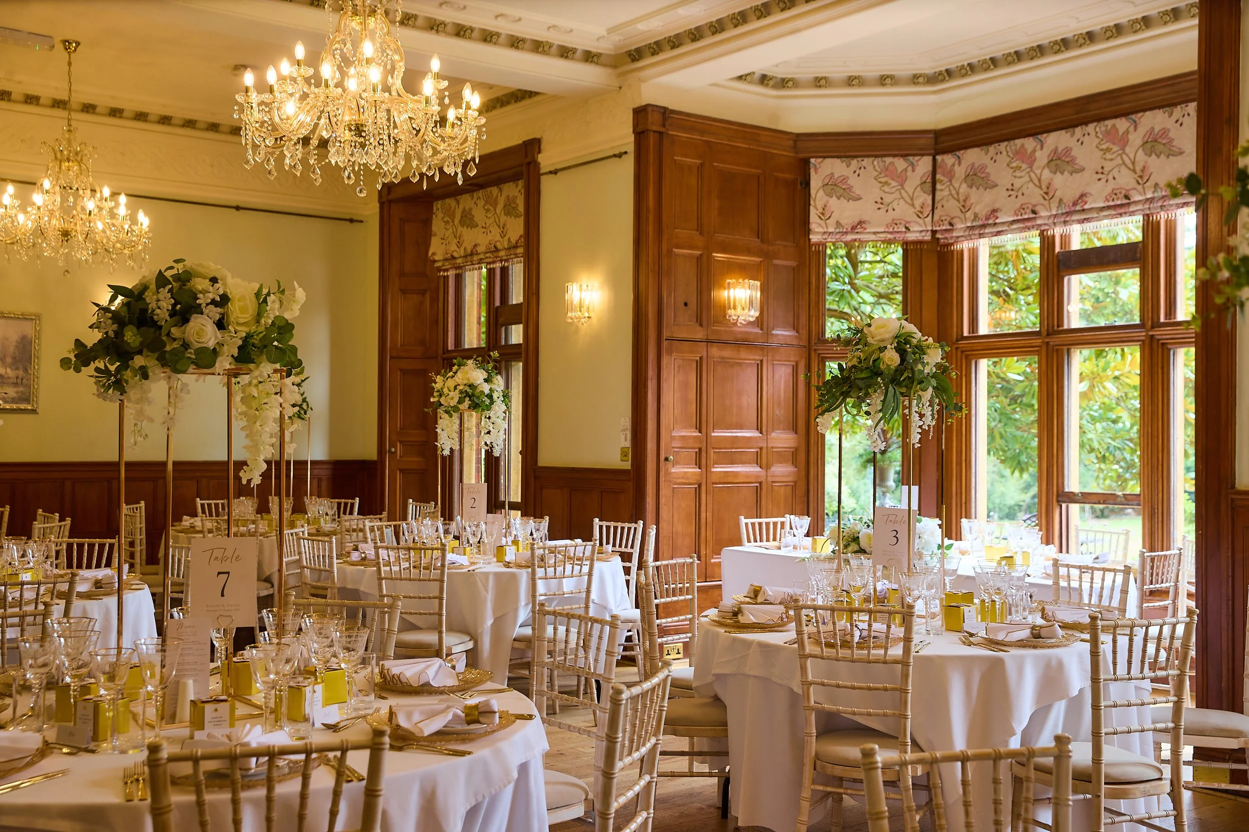 Elegant banquet hall decorated with chandeliers, large floral centerpieces, and white tablecloths, set up for a wedding or formal event.