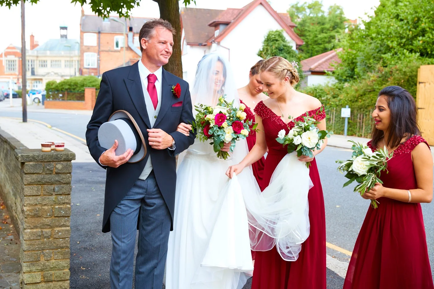 A bride and three bridesmaids, all holding bouquets, standing outside on a sidewalk near a street, with a man in a suit and hat holding a gray hat in front of them. The bride is wearing a white wedding dress and veil, while the bridesmaids are in red