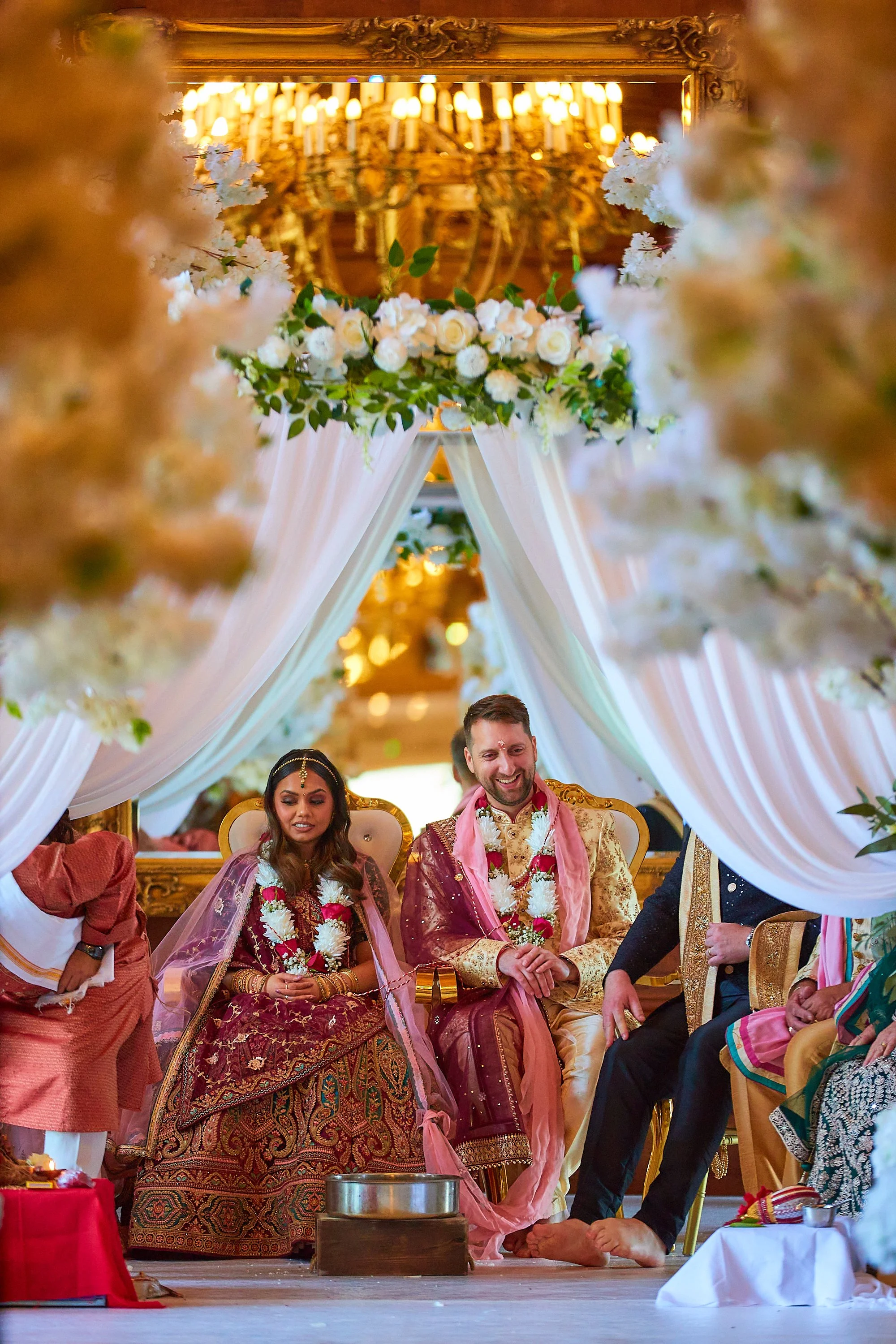 A couple dressed in traditional Indian wedding attire sitting on a decorated stage under a white canopy with floral arrangements, during a wedding ceremony.