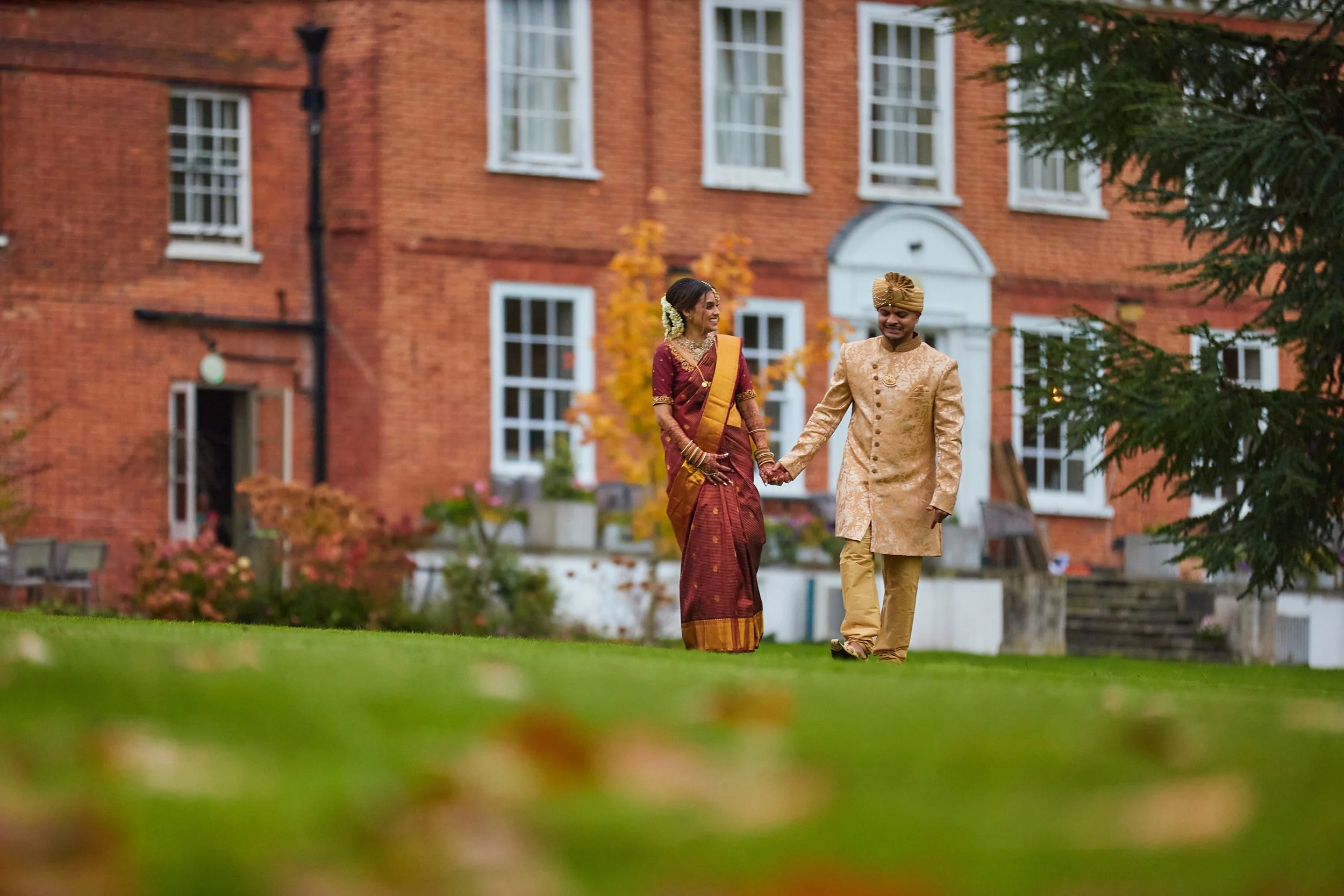 A couple in traditional Indian wedding attire holding hands and walking on a grassy area in front of a red brick building with white window frames. The woman is wearing a maroon and gold sari with jewelry, and the man is wearing a beige sherwani with